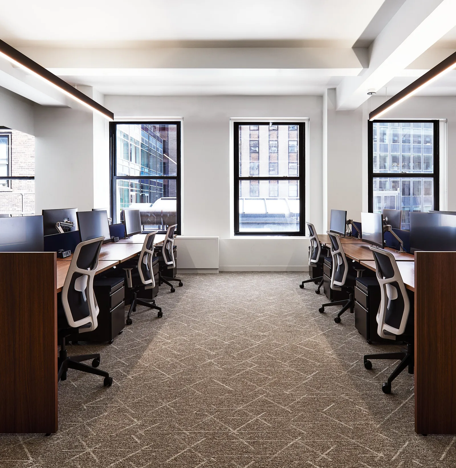 aisle between desk clusters toward three black-framed windows, chairs and lineal lights overhead