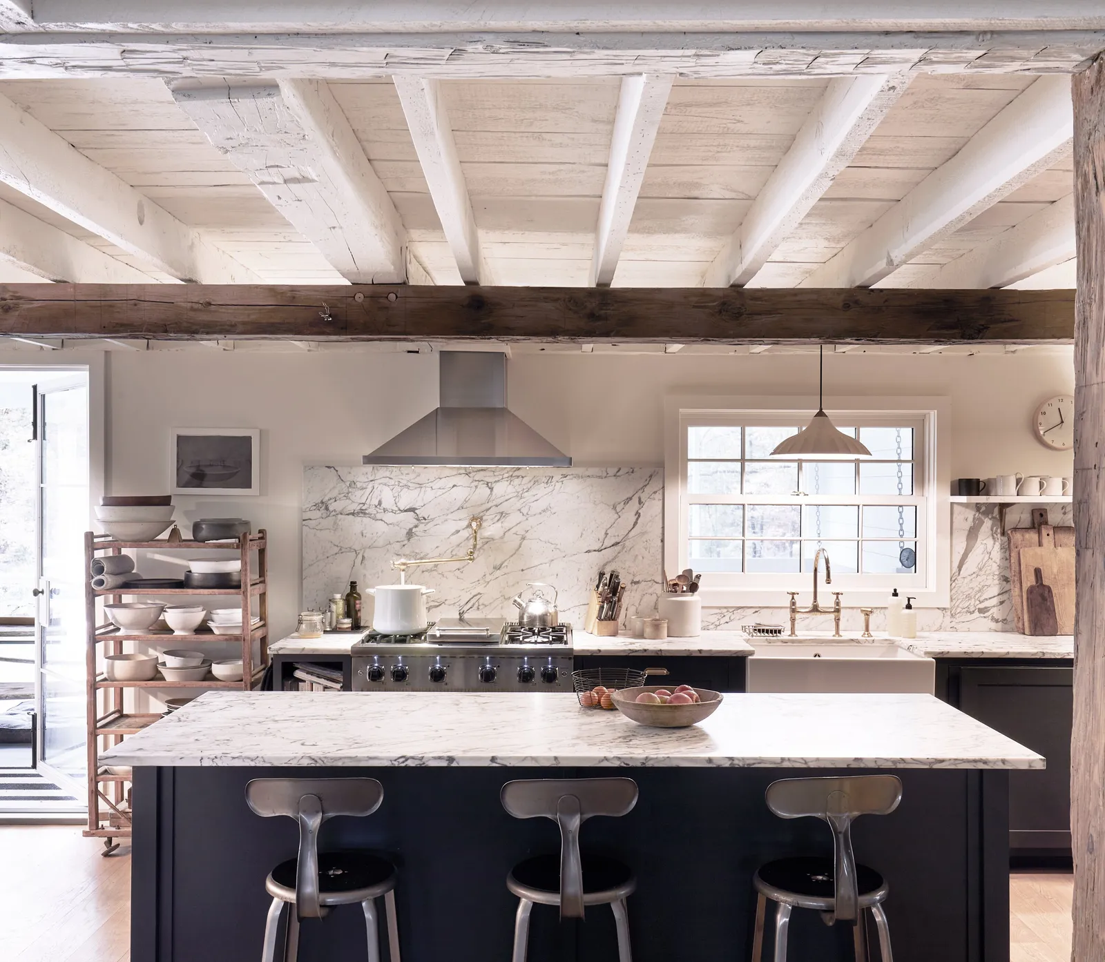 kitchen with a marble island, three metal stools, a range hood and whitewashed ceiling beams