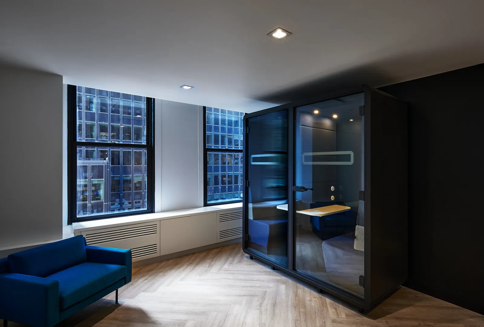 corner lounge with blue sofa, tall glass phone booth, radiator cover and windows on herringbone floor