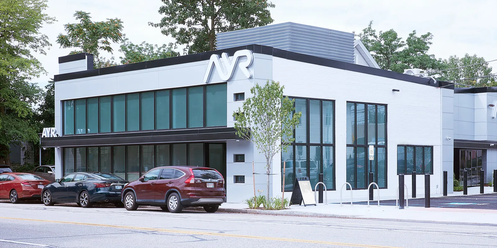 corner view of the white and glass building with the AYR logo, a young tree, and cars along the street