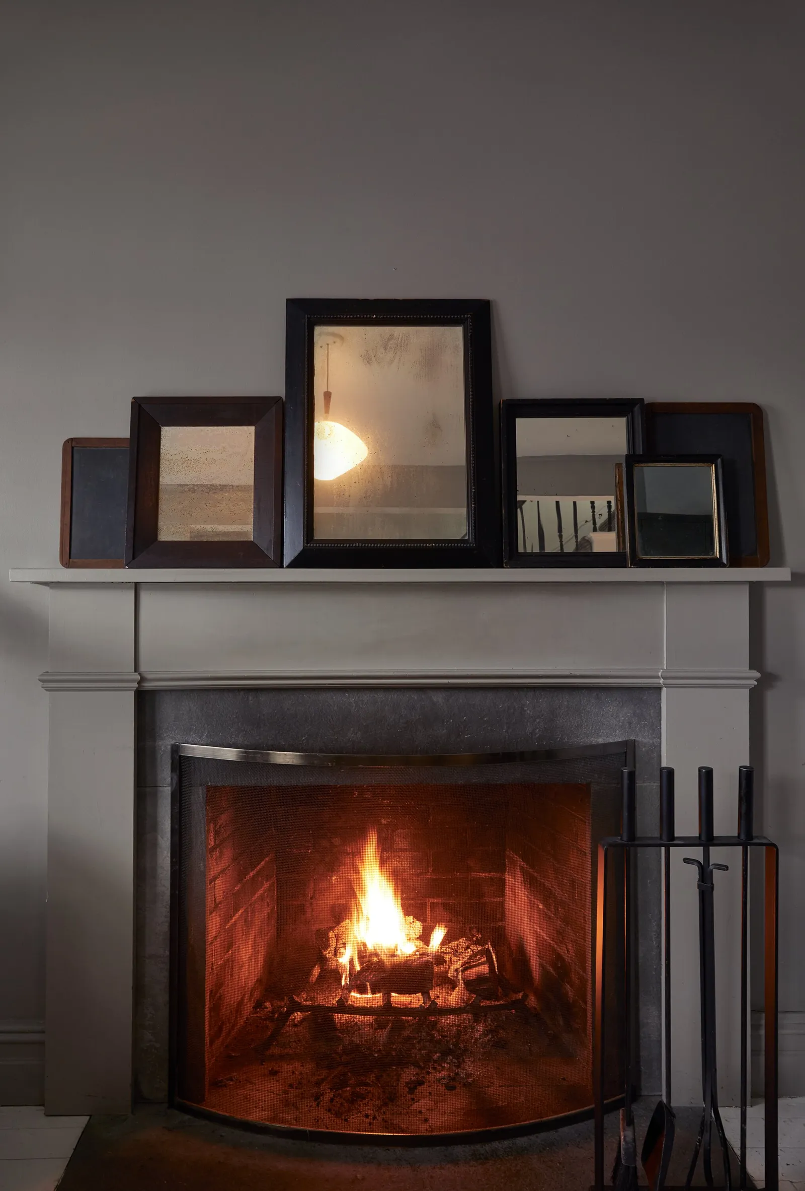 fireplace with a curved screen and a lit fire, framed mirrors on the mantel and fire tools at the side