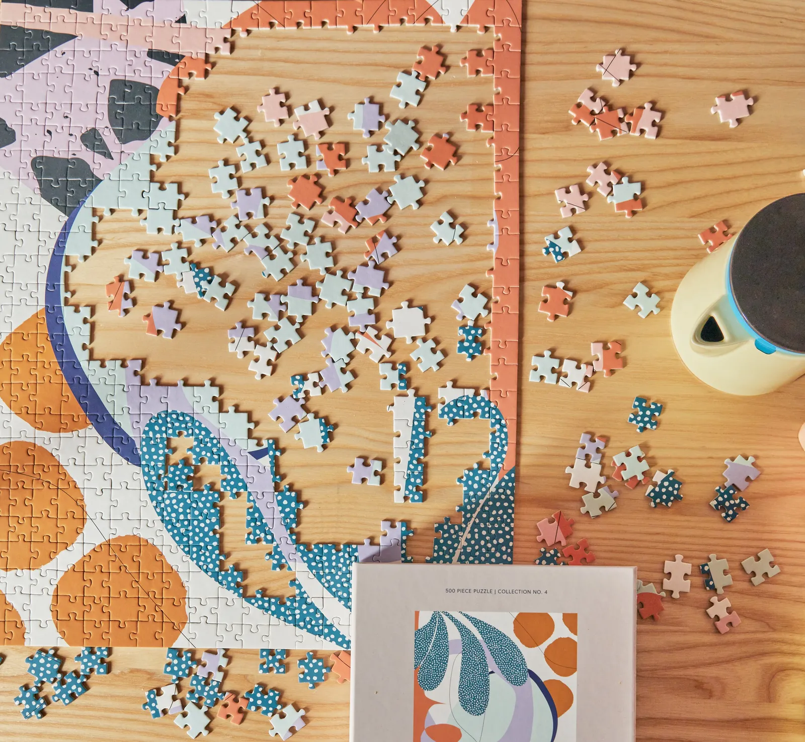 overhead view of a half-finished jigsaw puzzle on a wood table, loose pieces, kettle and puzzle box