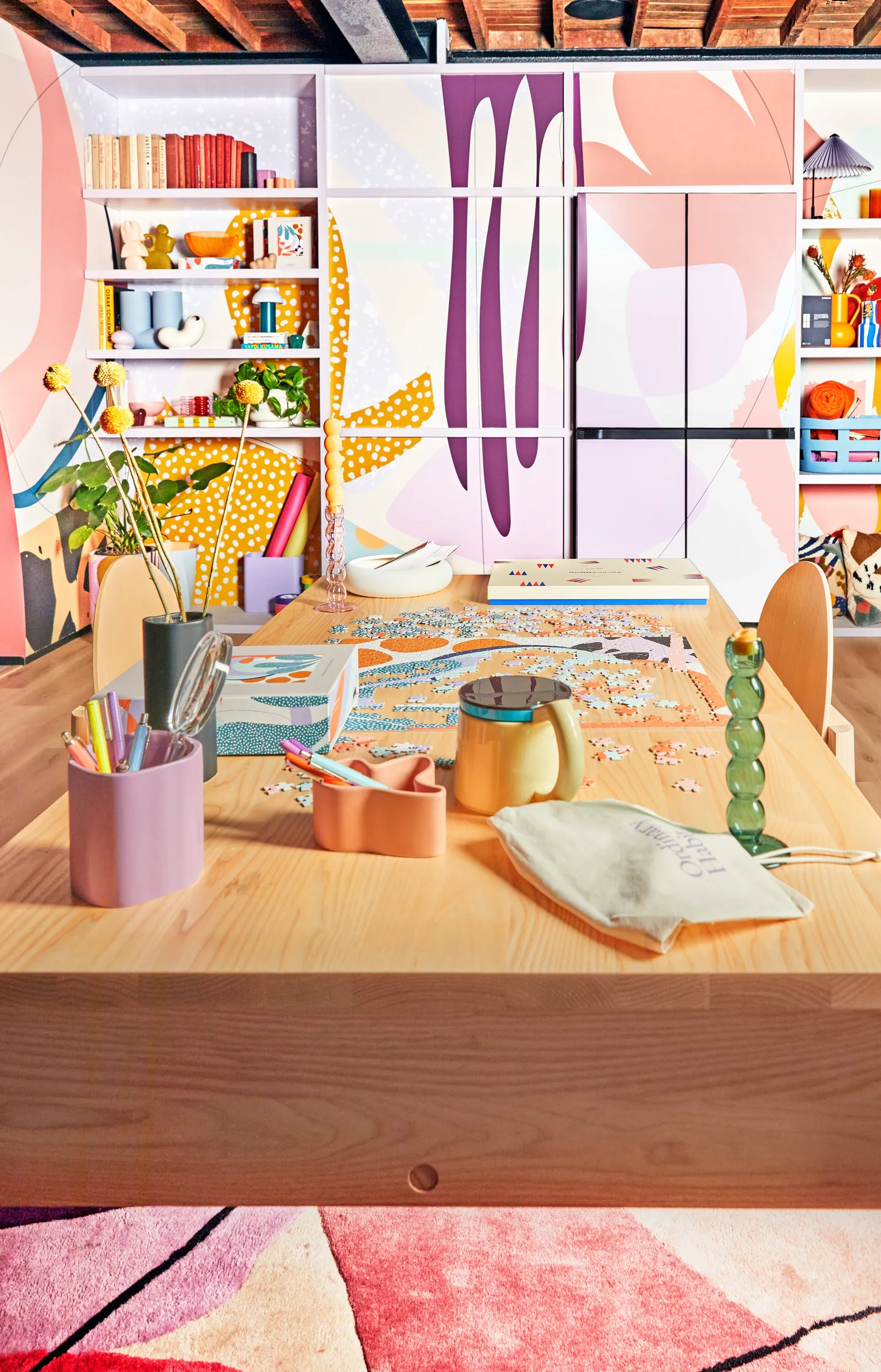 wood table with pencil cups, puzzle and kettle in front of the mural wall of shelves and refrigerator