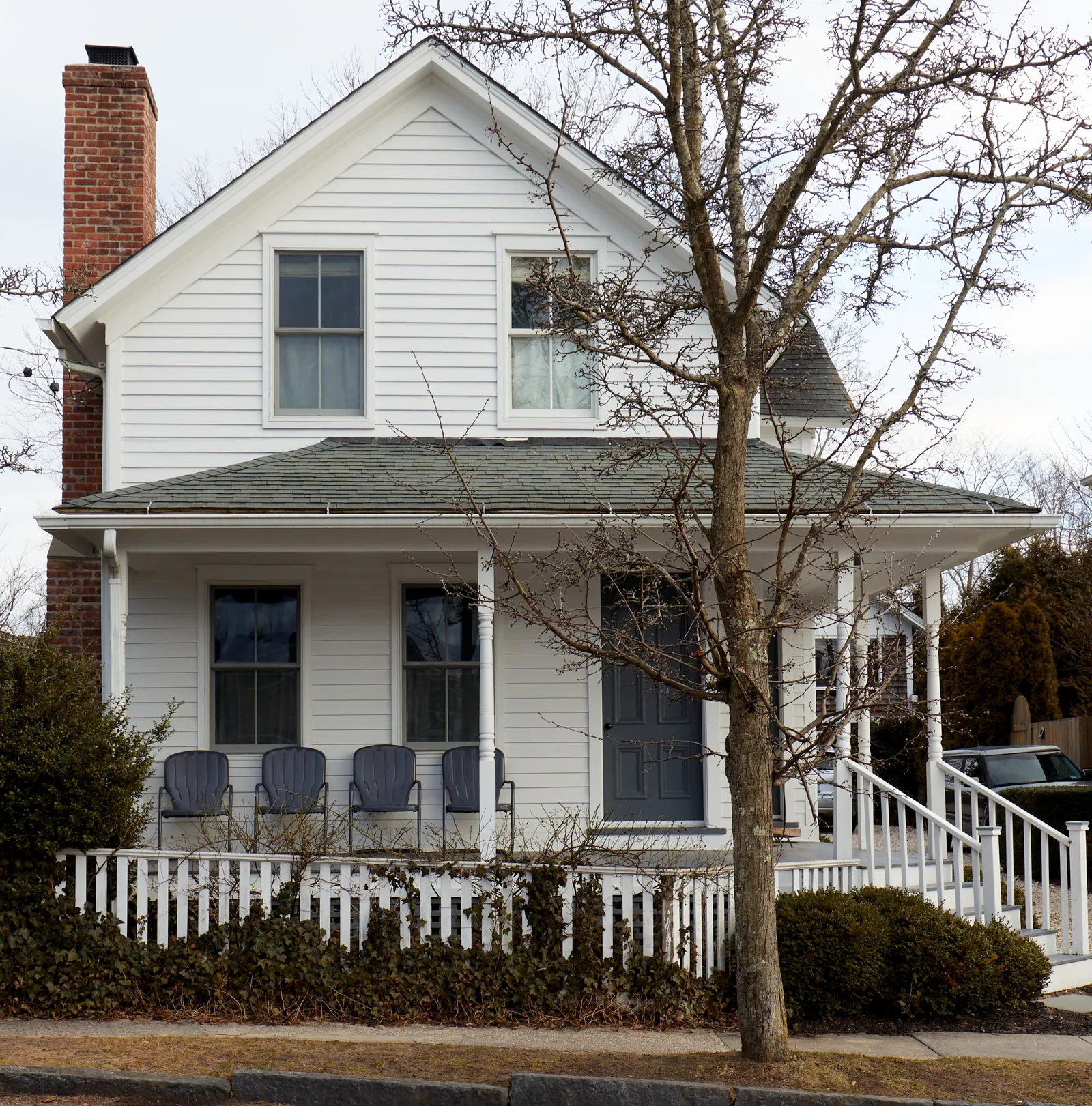 front of the white clapboard house with a porch, blue chairs, a white picket fence and a bare tree