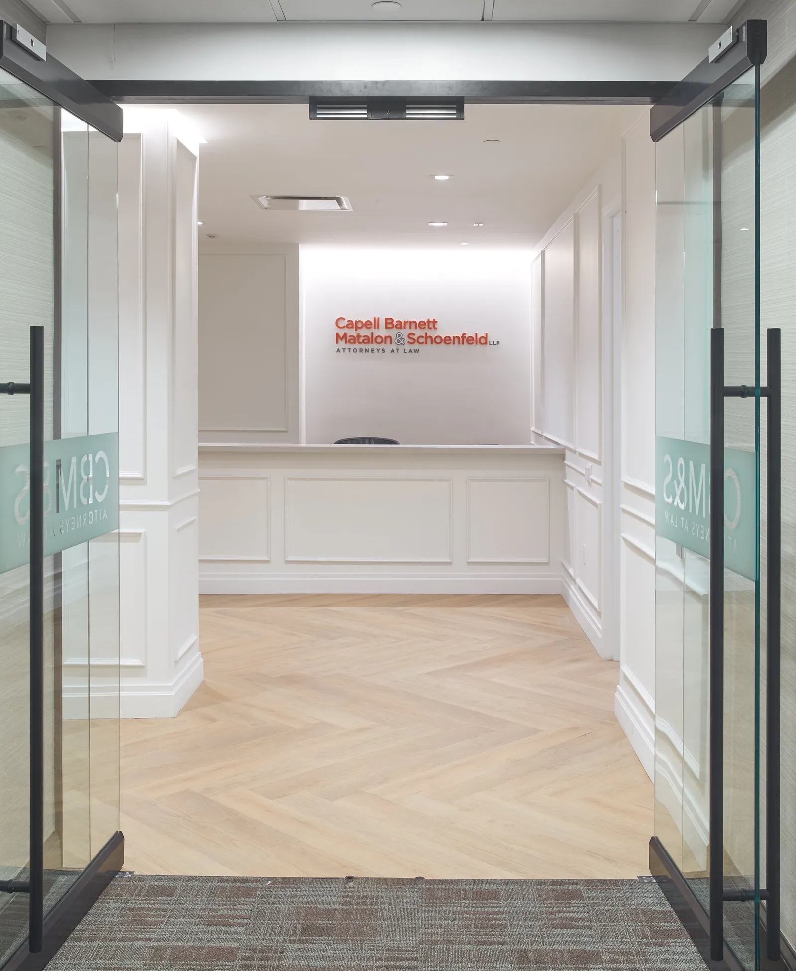 entry through open glass doors toward reception, red firm name on the wall and herringbone floor