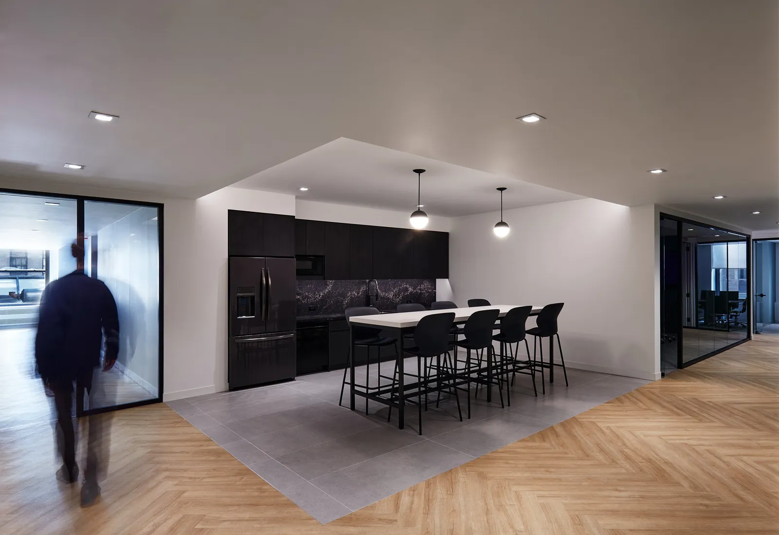 same kitchen with a blurred man in a dark jacket walking past the glass wall at left