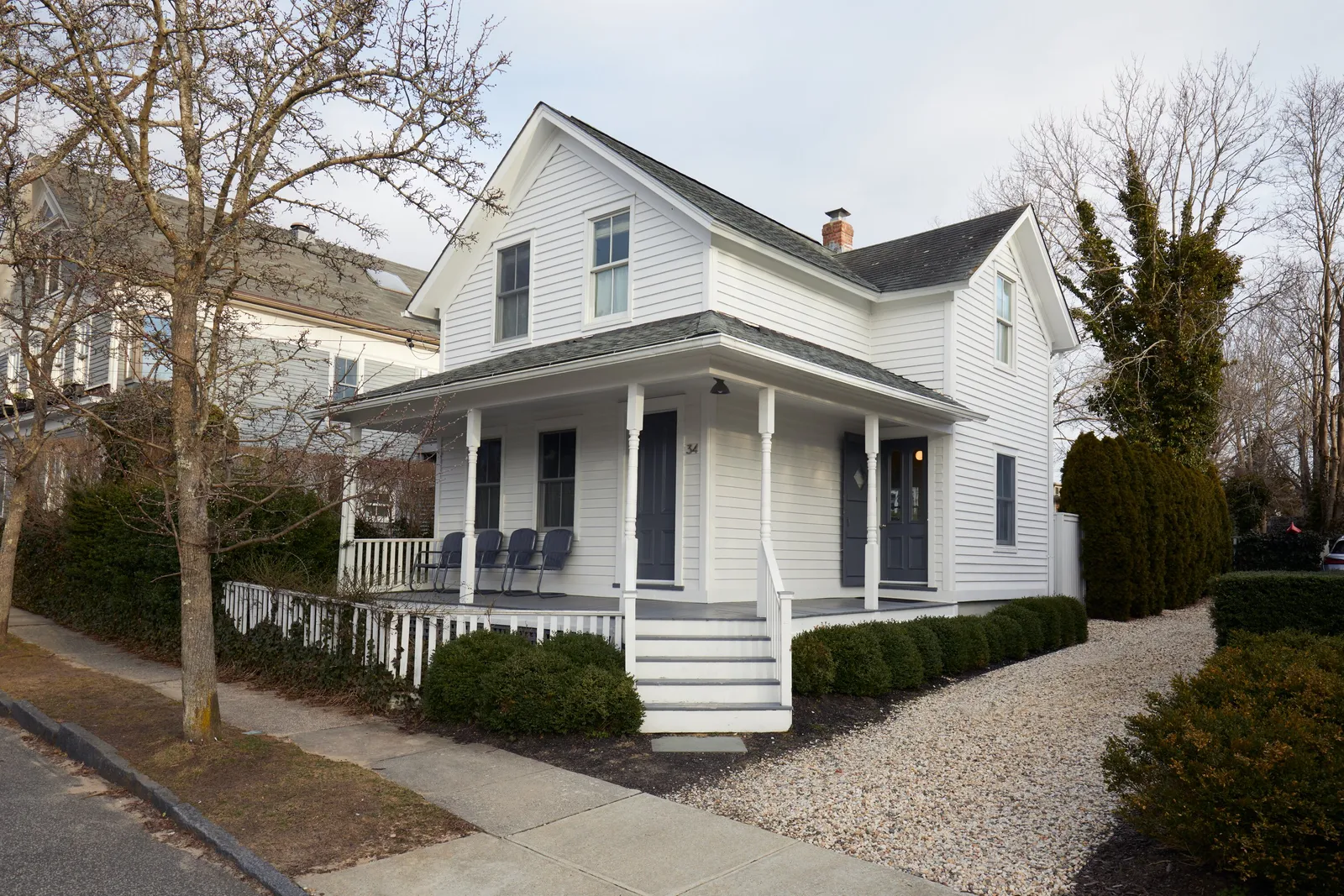 white house seen from the street at an angle, with a gabled porch, gravel drive and clipped hedges