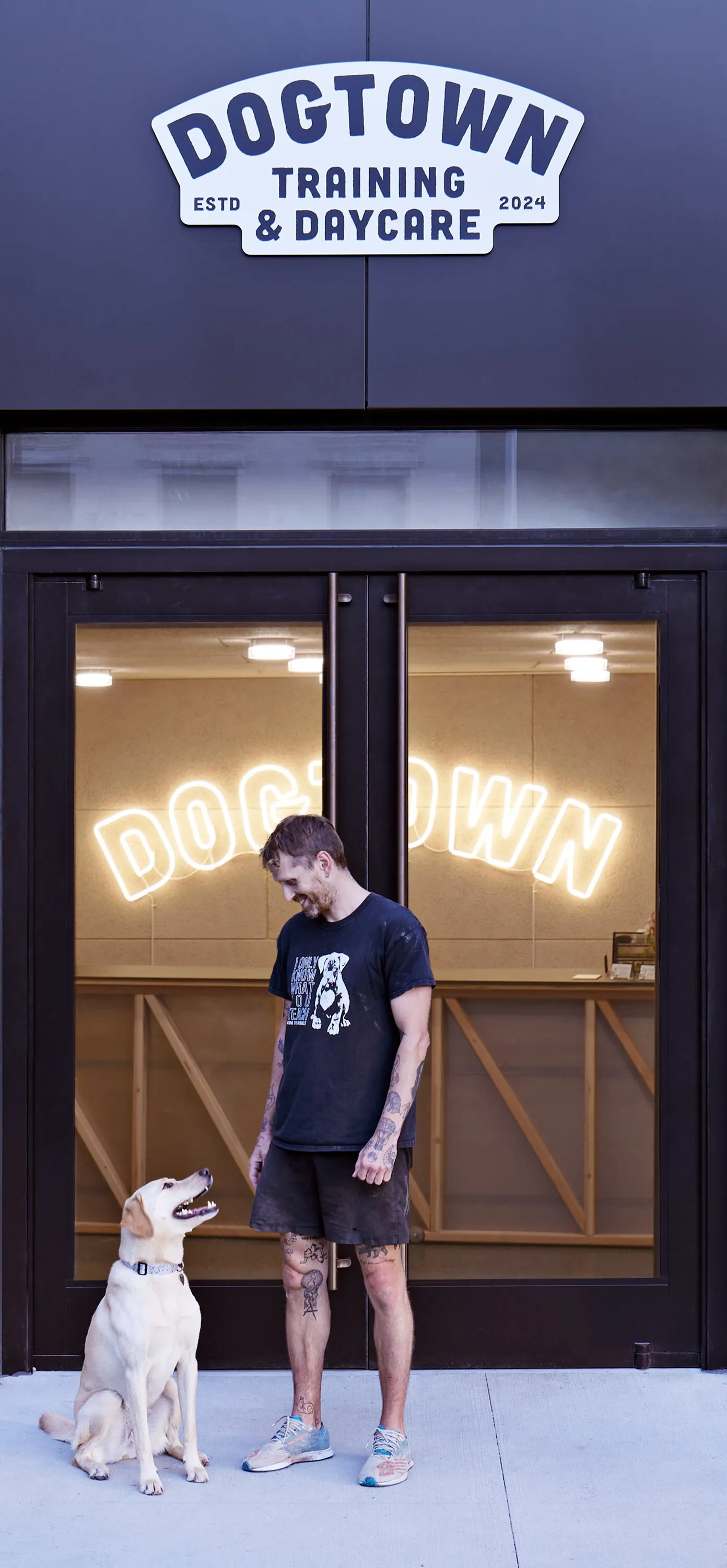 man with tattoos and a light-colored dog sitting outside the glass doors, the DOGTOWN Training and Daycare sign above