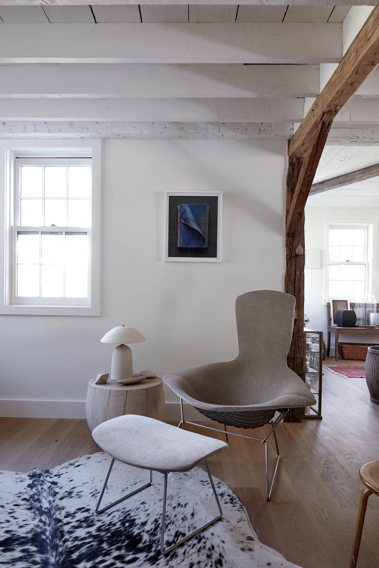 modern wire lounge chair with ottoman on a cowhide rug beside a window, with a rough timber post and a blue artwork