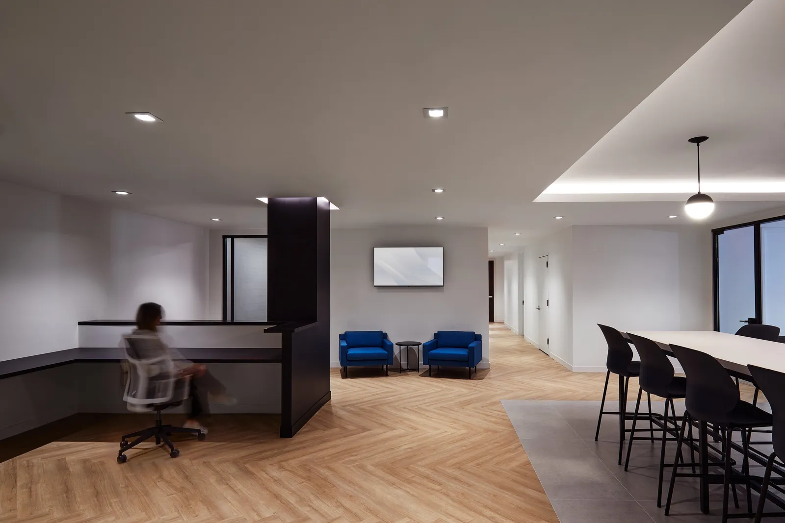 reception area with a seated blurred person at a black desk, two blue armchairs and a hallway beyond