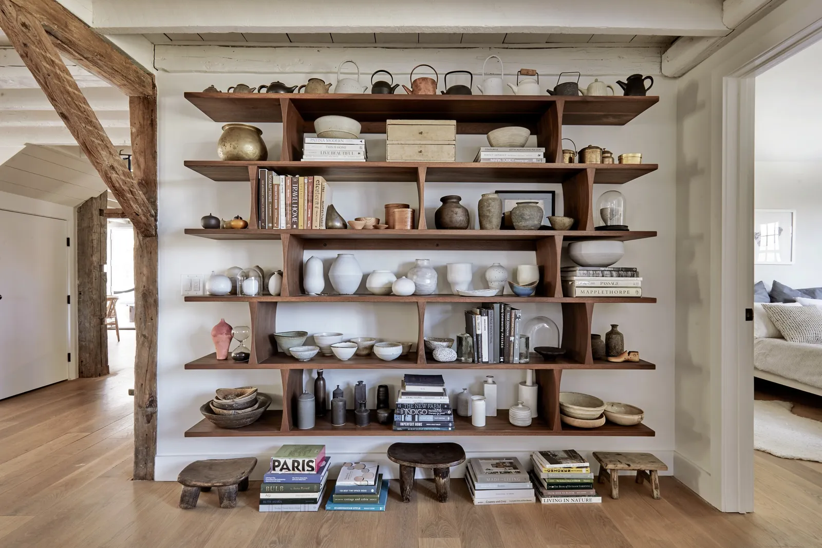 open wooden shelving of ceramics, teapots and books against a white wall between timber posts