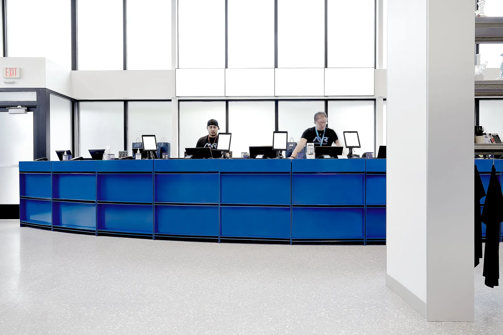 two staff at computers behind the long blue counter, with a white column and hanging clothes at right