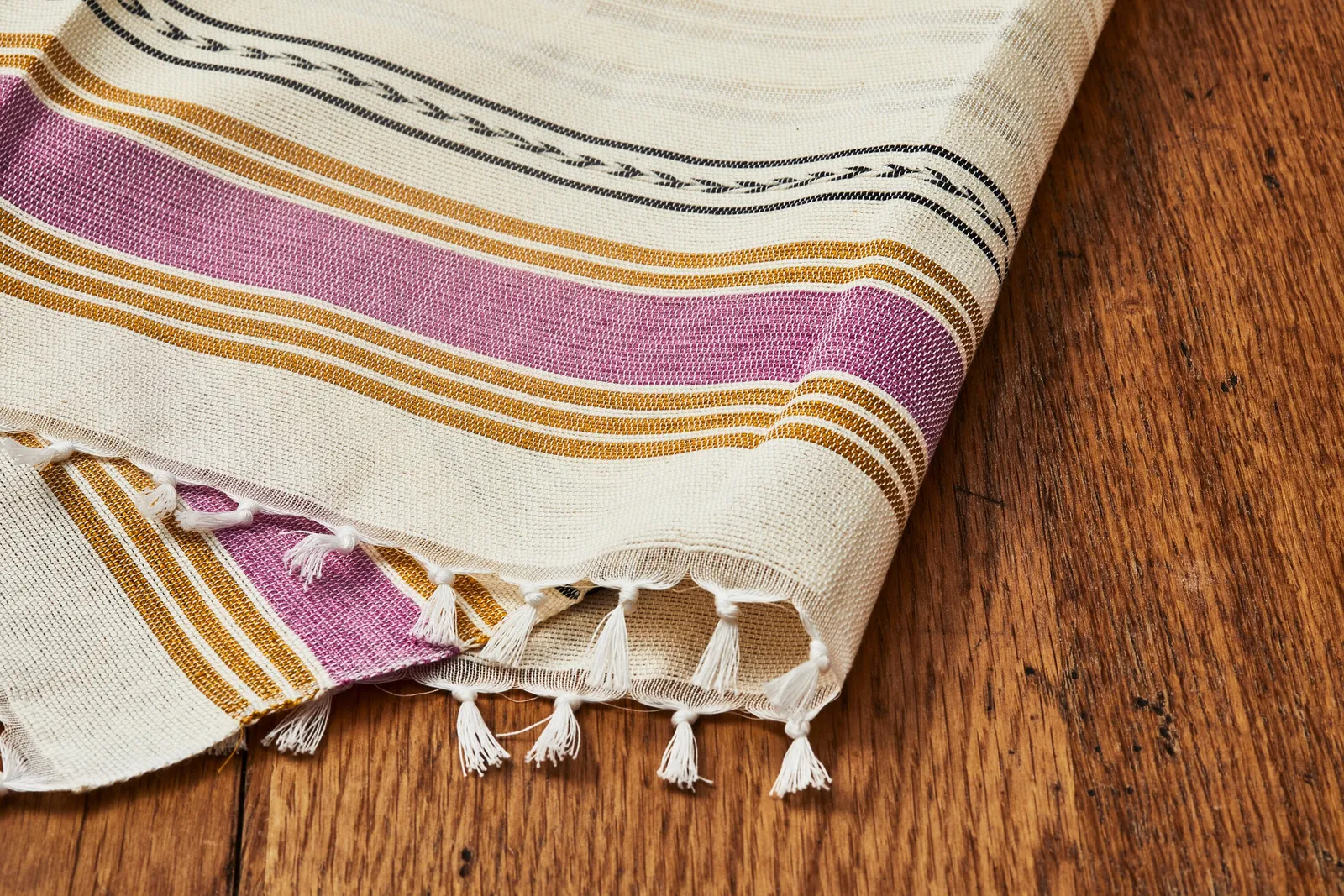 folded cream cloth with wide mauve and gold stripes, a woven black border and white tassels on a wood floor