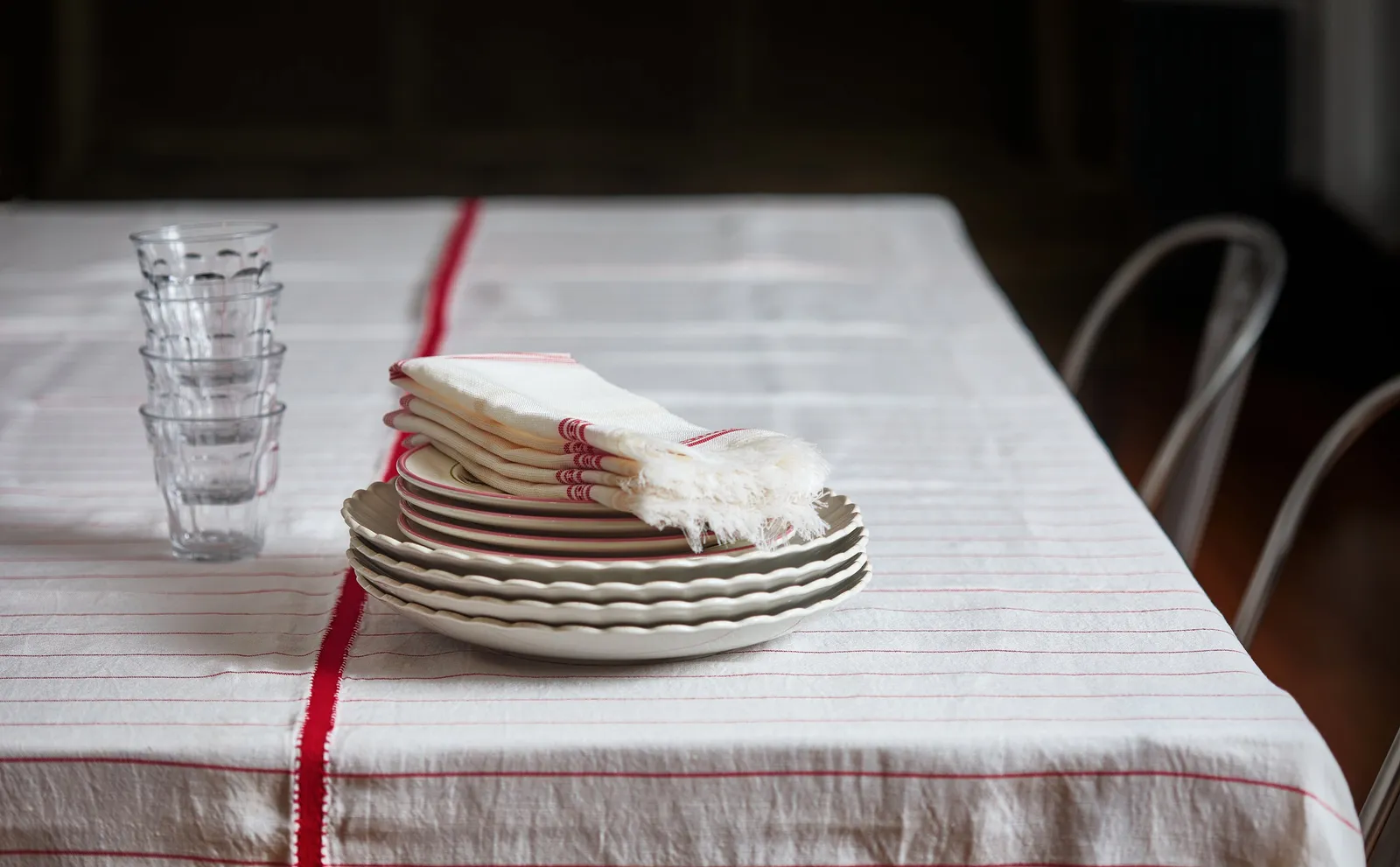 stack of plates with a folded fringed napkin and a stack of glasses on a cream tablecloth with a red center stripe, chairs behind