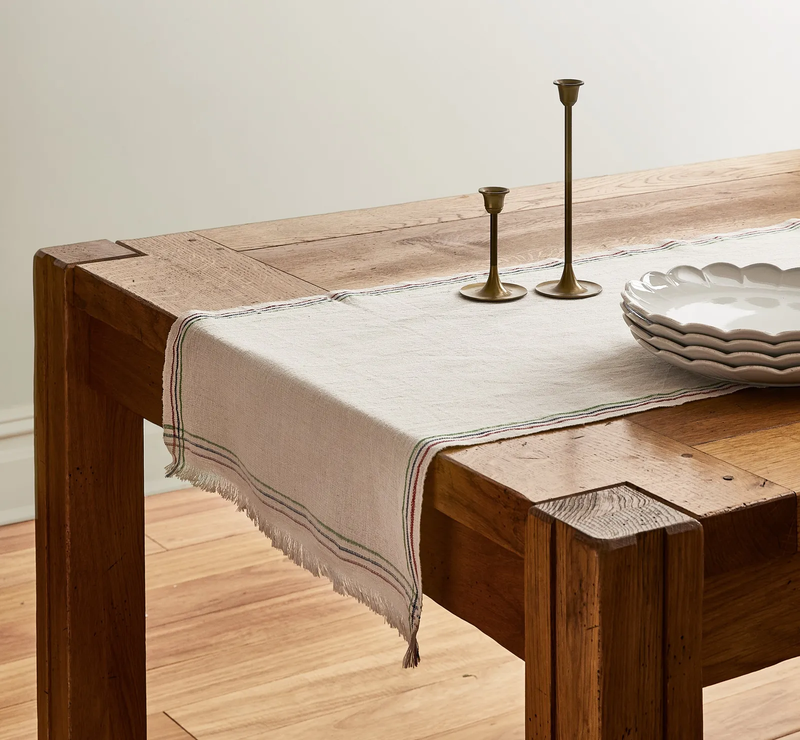 cream table runner with thin colored stripes draped over a heavy wood table, with two brass candlesticks and scalloped plates