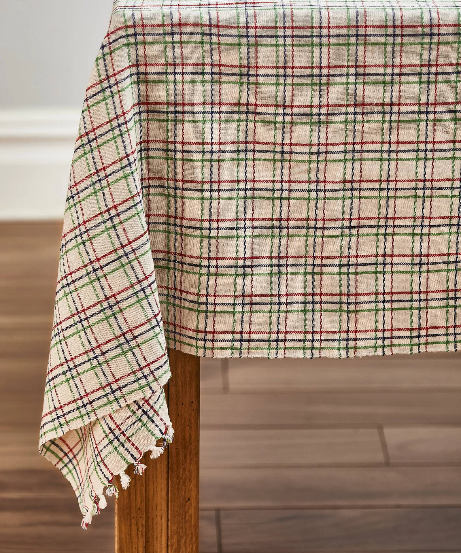 red, green and navy plaid tablecloth hanging over a wood table corner, with frayed edges and small tassels on a dark floor