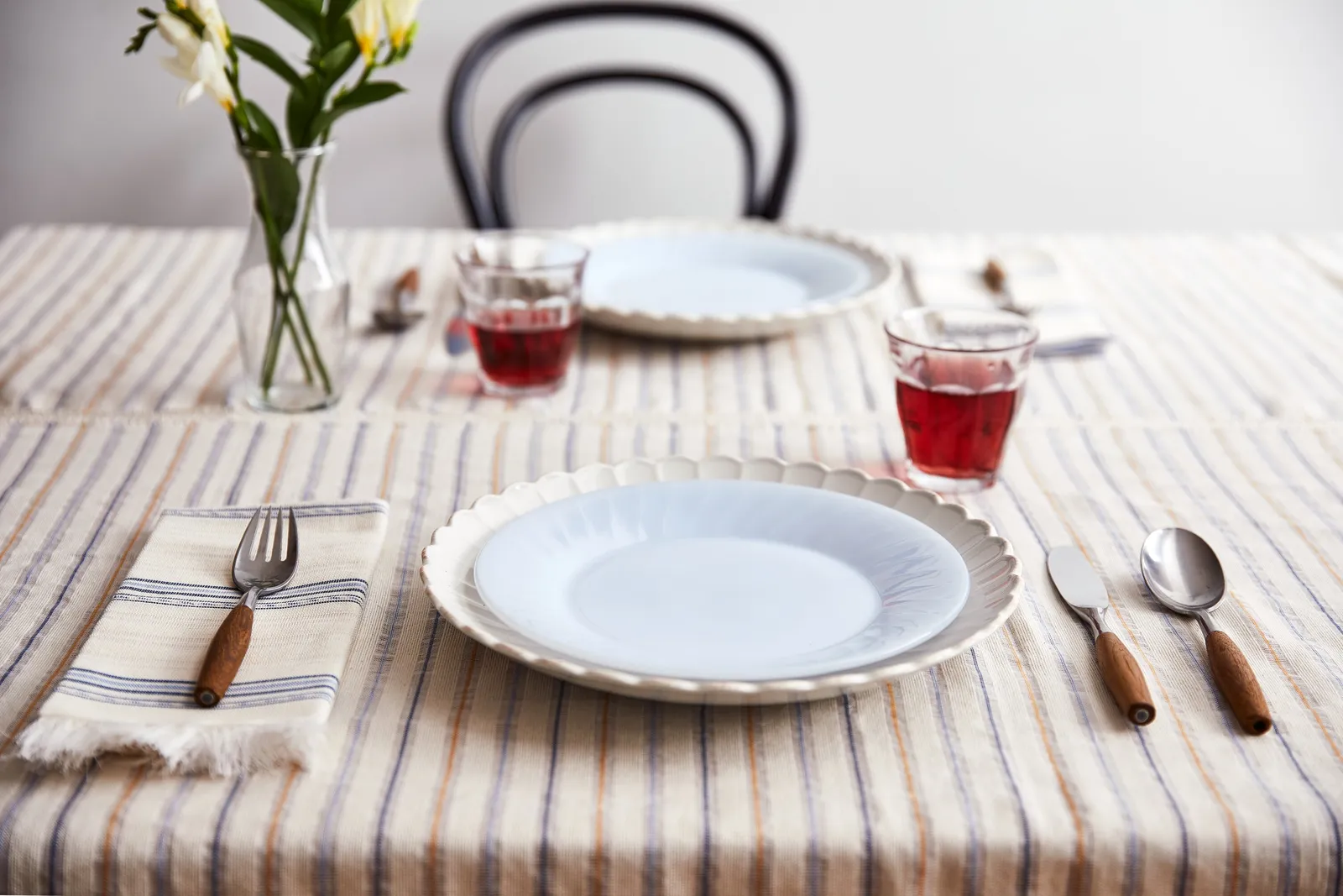 table set on a striped cream cloth with scalloped white plates, napkins, wood-handled cutlery, red drinks and flowers, a bentwood chair behind