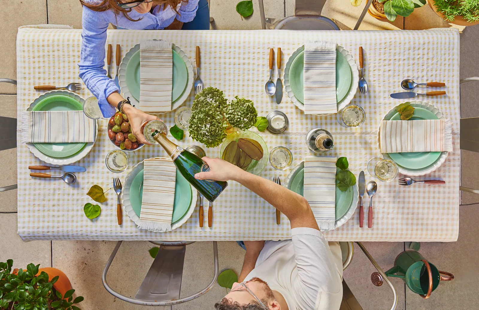 overhead view of two people pouring champagne at a long table with a pale yellow gingham cloth, green plates and striped napkins