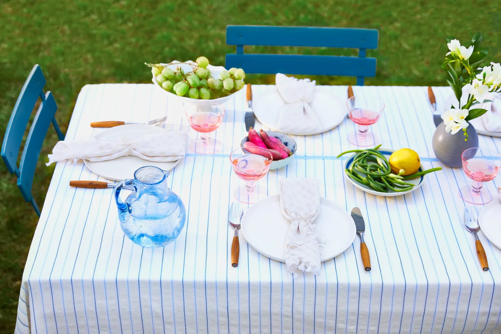 outdoor table on grass with a white cloth striped in blue, pink drinks, grapes, a blue glass pitcher, flowers and blue chairs