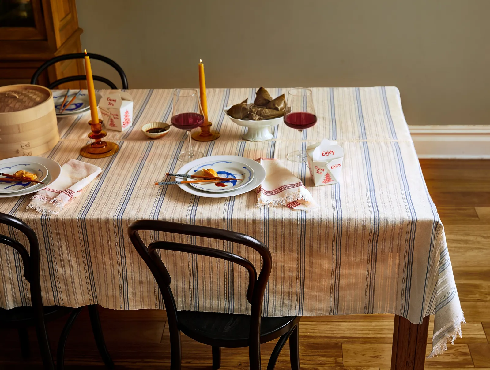 dinner table with a cream and blue striped cloth, yellow candles, wine, takeout boxes and plates with chopsticks, black bentwood chairs
