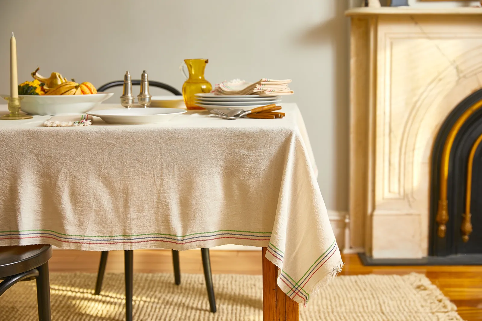 cream tablecloth with green and red edge stripes on a table with candle, plates, gourds and a yellow pitcher, beside a marble fireplace