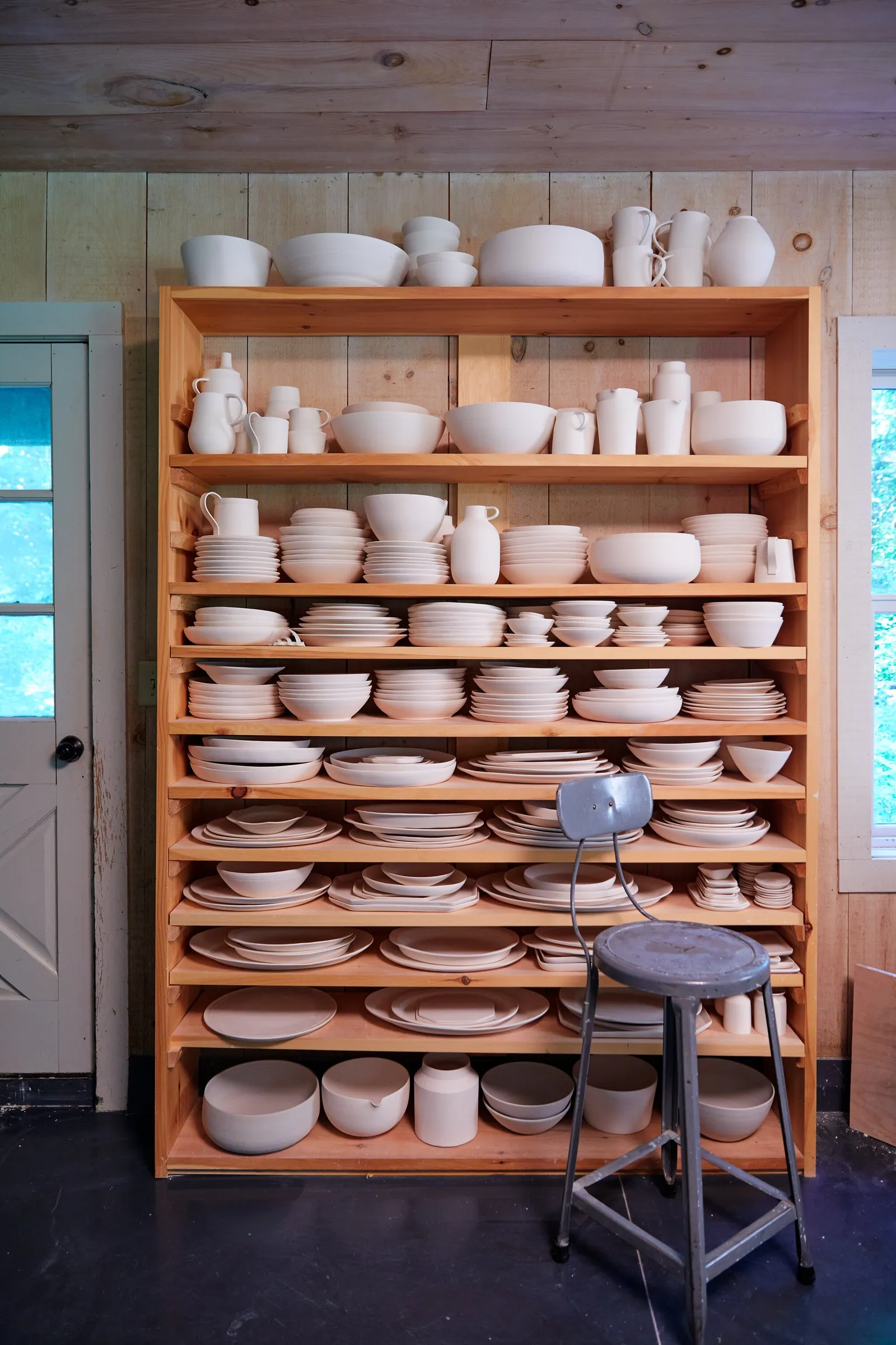 a wooden shelving unit filled with rows of white and cream ceramic bowls, plates and jugs in a bright studio