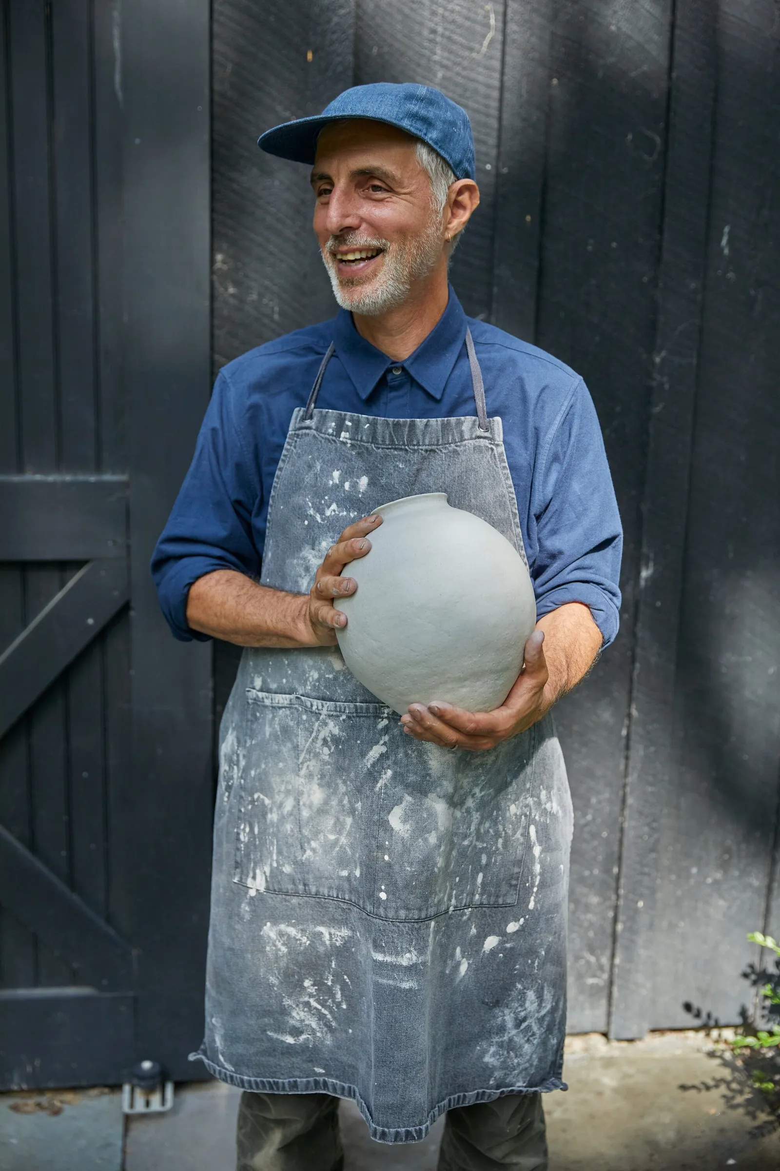 a smiling man in a denim cap and paint-splattered apron holding a pale round ceramic vase in front of a dark barn wall