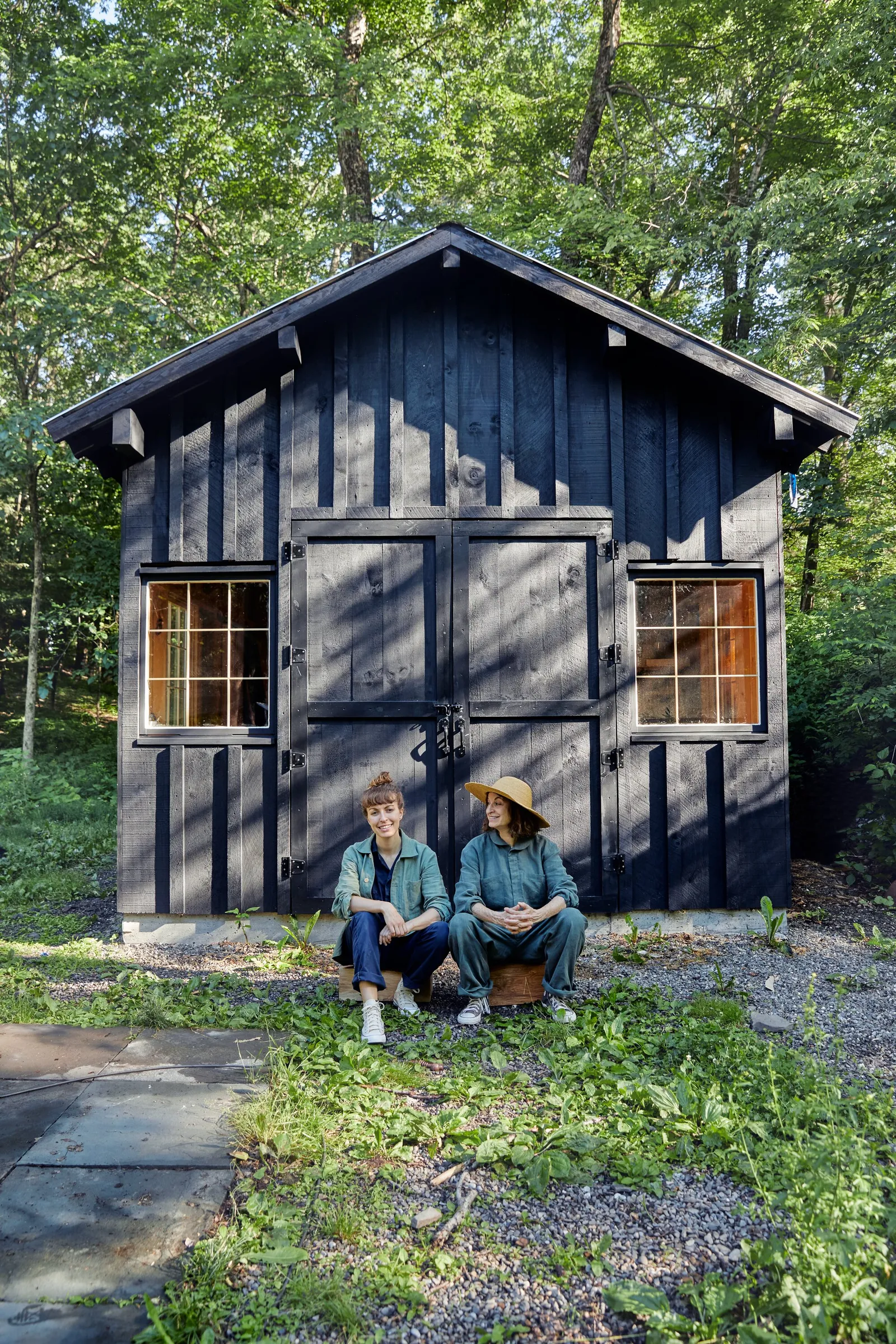 two people in work clothes sitting side by side in front of a dark wood barn with double doors, surrounded by trees