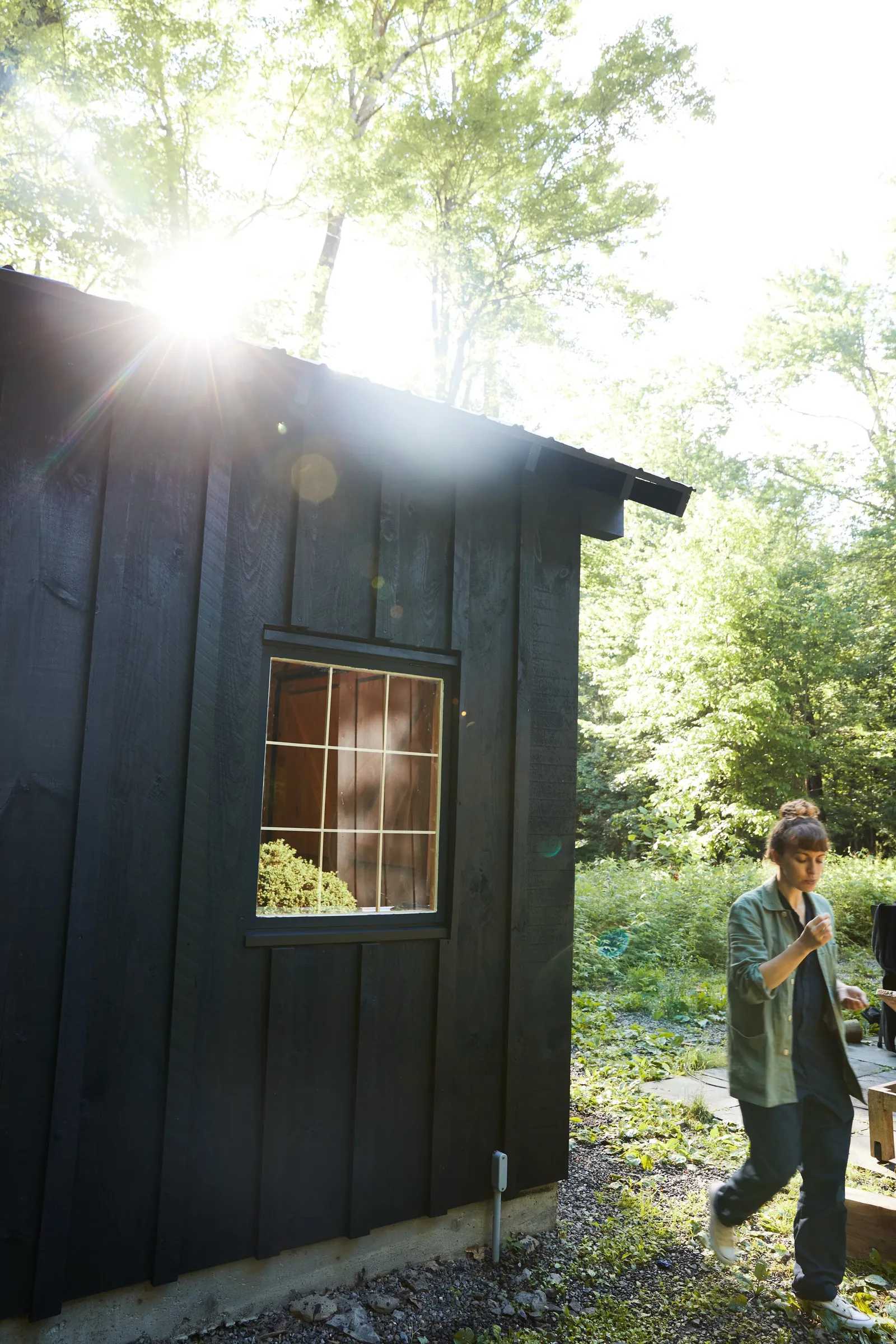 sunlight flaring over the roof of a dark wood barn as a woman in a green work jacket walks past in the garden