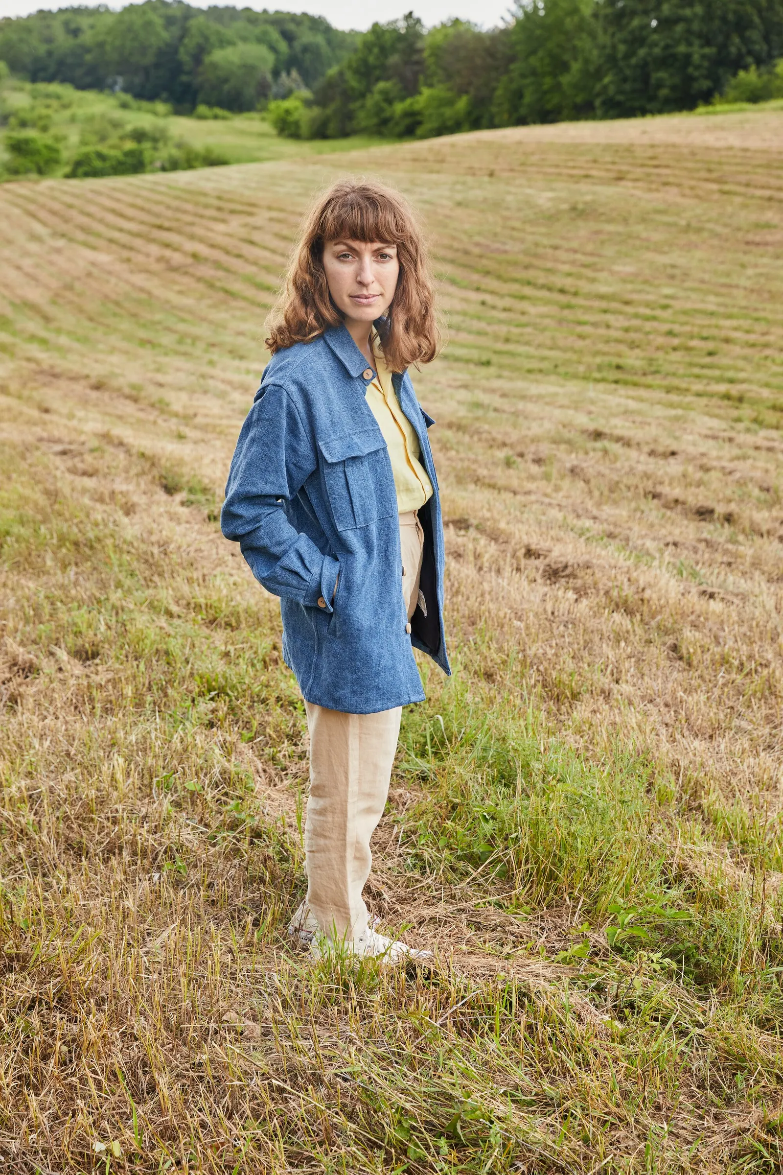a woman in a blue denim shirt jacket and khaki pants standing in a mown golden field