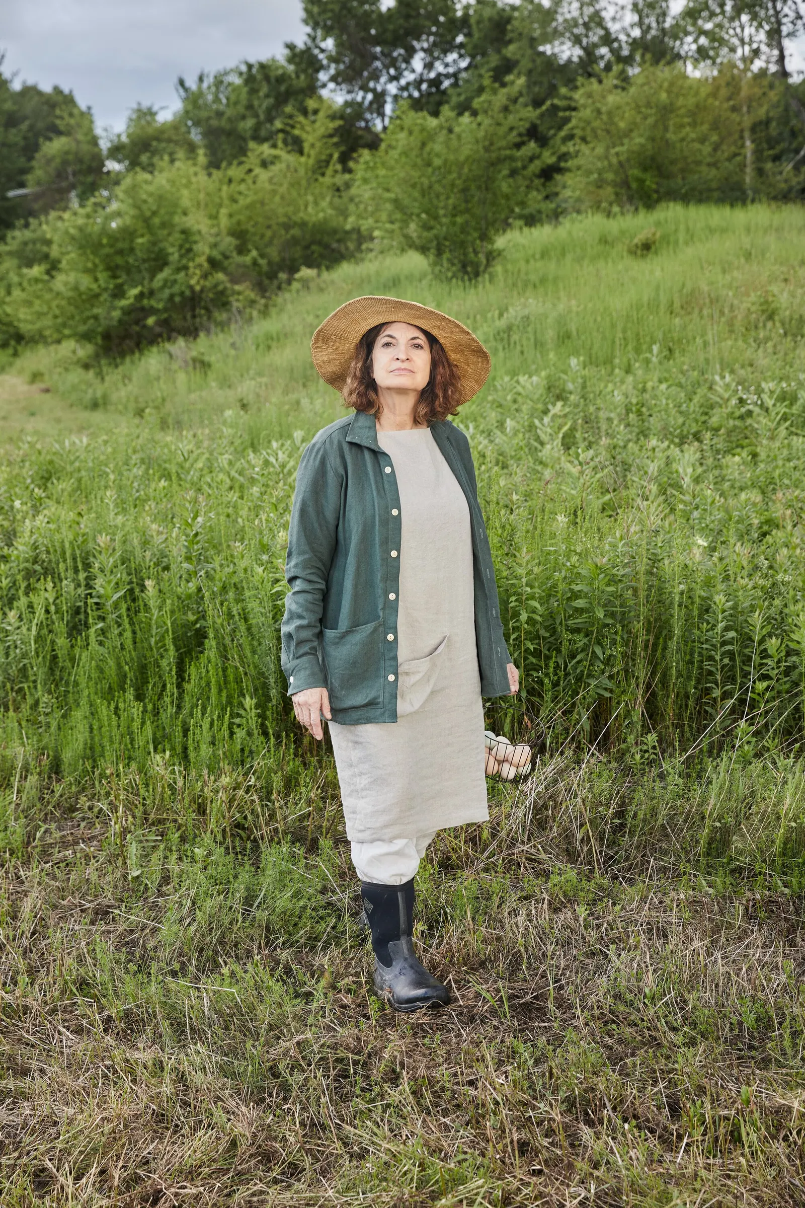 a woman in a straw hat, green jacket and linen dress standing in tall green grass, looking up