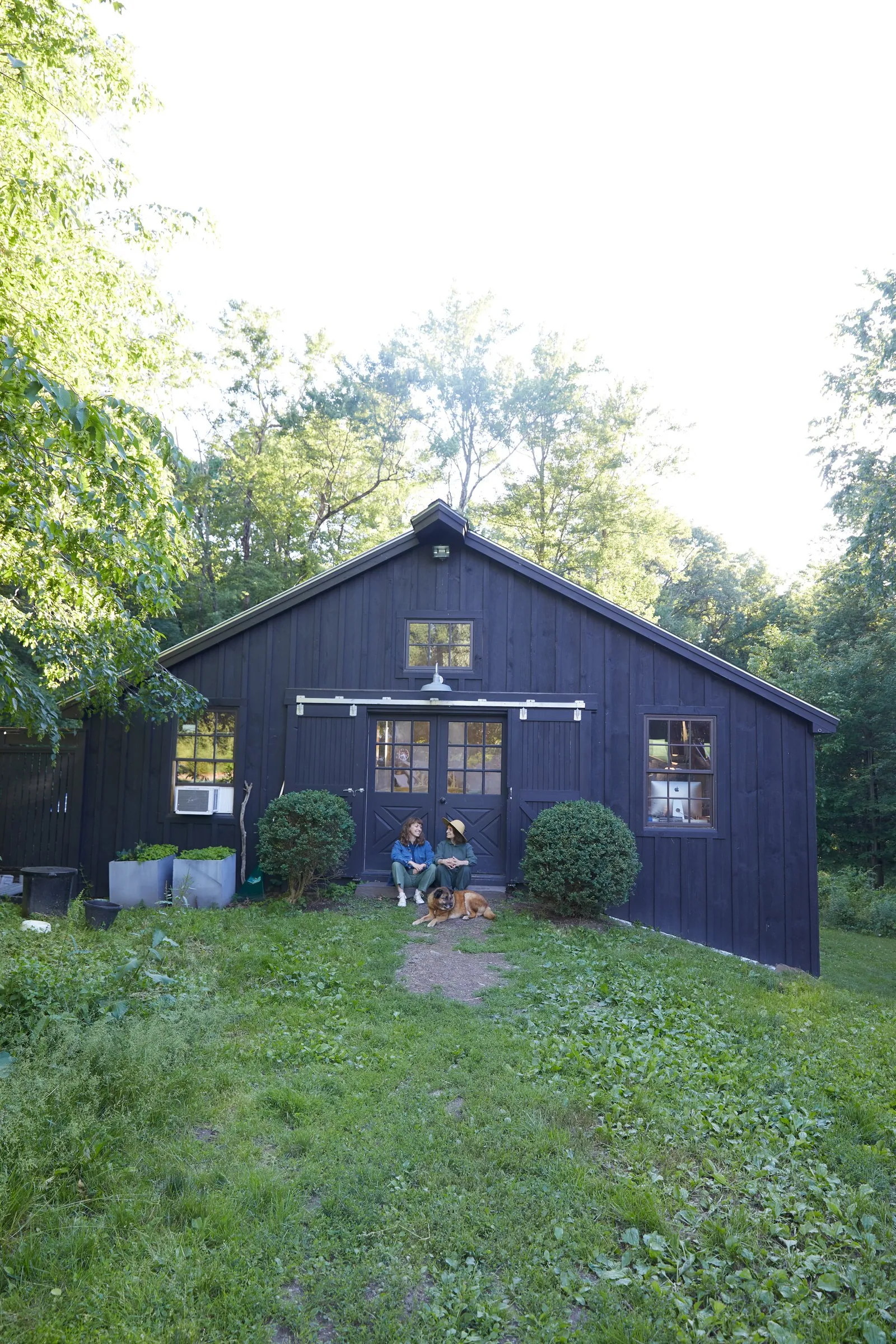 a dark barn seen from across a green lawn, with two people and a dog sitting small by the doors