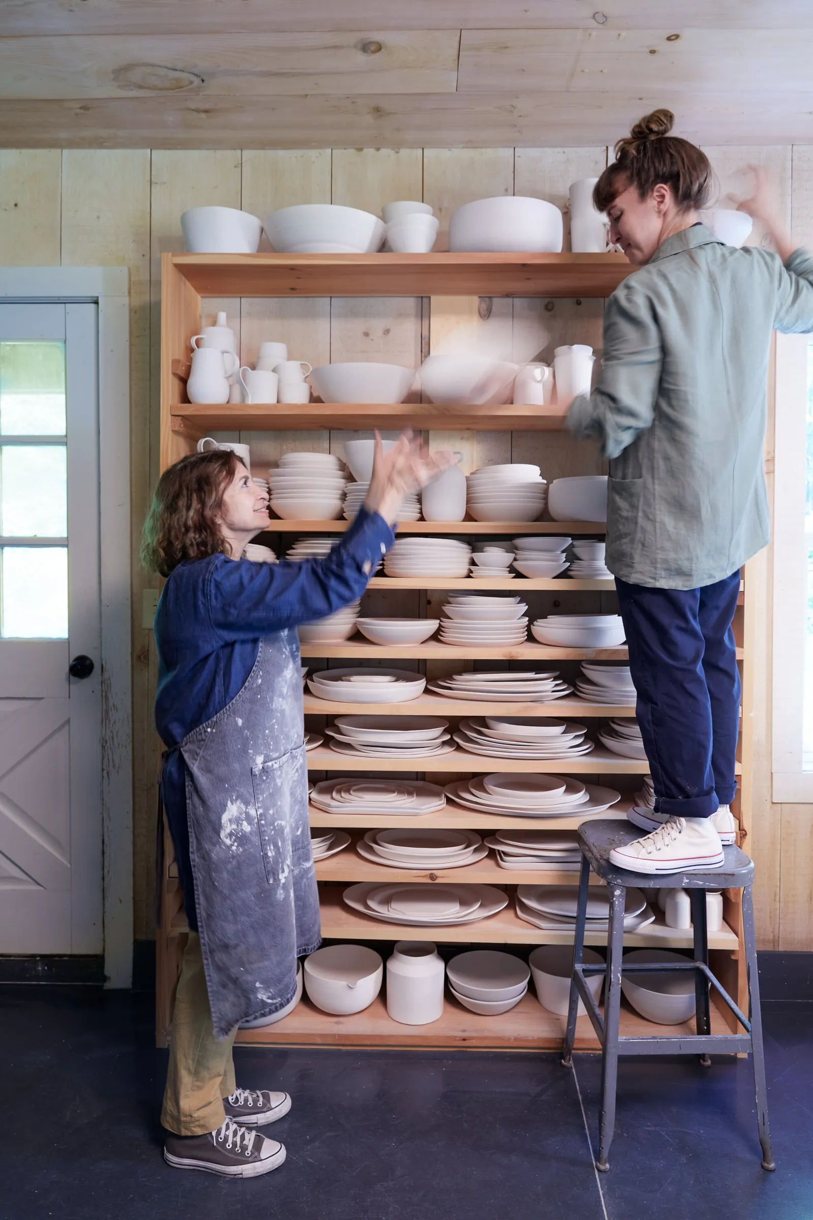 a woman in a denim apron reaching toward a shelf of ceramics beside another person standing on a step stool
