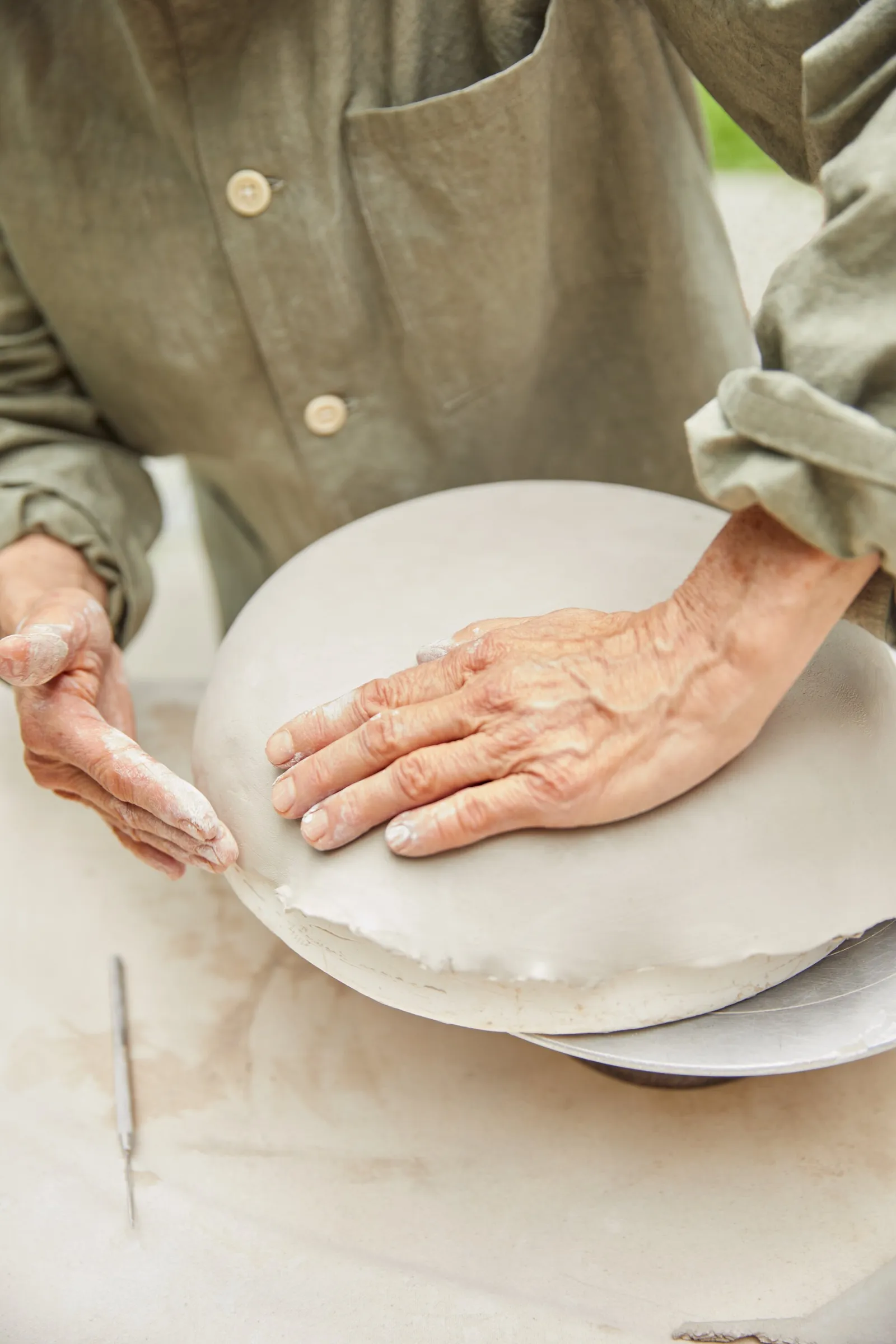 hands pressing down on a gray clay plate being shaped, with a sleeve of an olive green work shirt visible