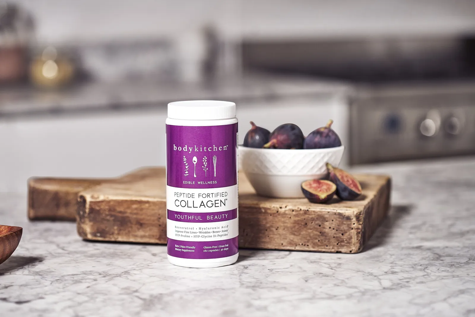 purple collagen jar on a marble counter before a wood board holding a white bowl of figs, with a blurred stove behind