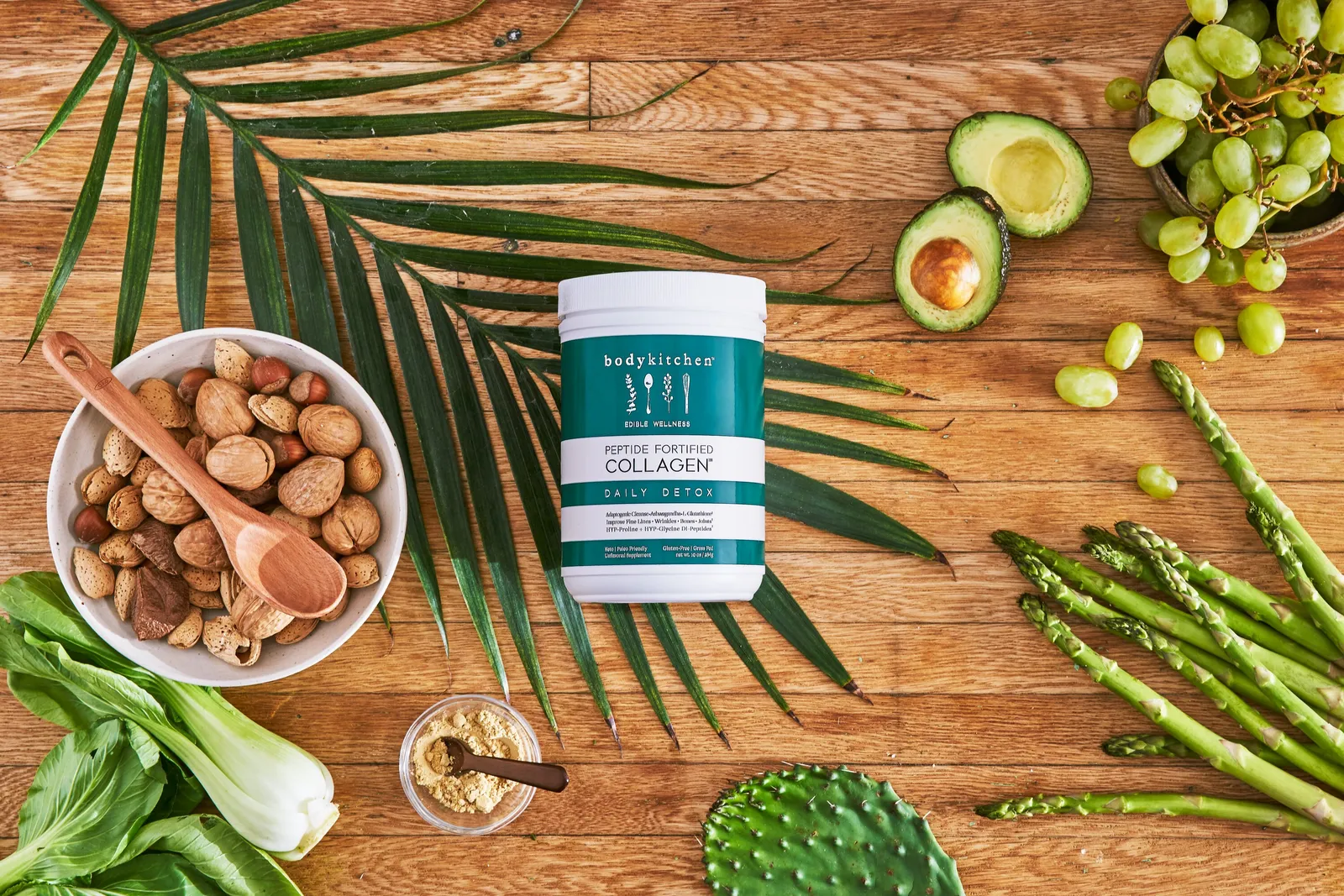 overhead view of a teal collagen jar on a wood table with a palm leaf, bowl of nuts, bok choy, asparagus, green grapes, avocado and cactus paddle