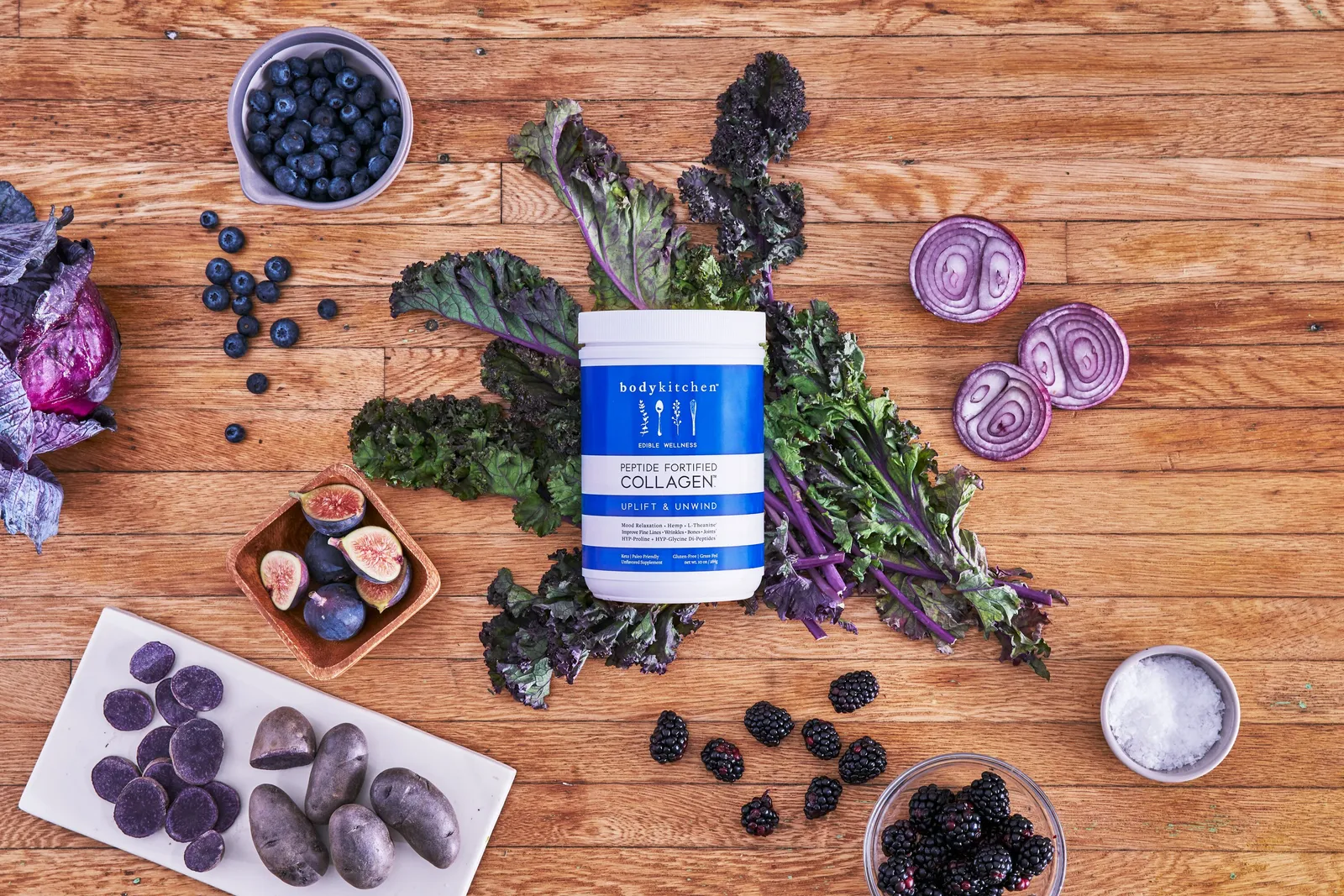 overhead view of a blue collagen jar on a wood table among purple kale, blueberries, blackberries, figs, purple potatoes and sliced red onion
