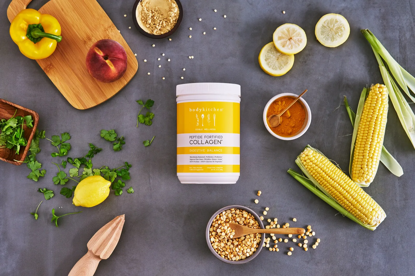 overhead view of a yellow collagen jar on a gray surface with corn on the cob, lemons, a peach, parsley, a yellow pepper, turmeric and a wooden juicer