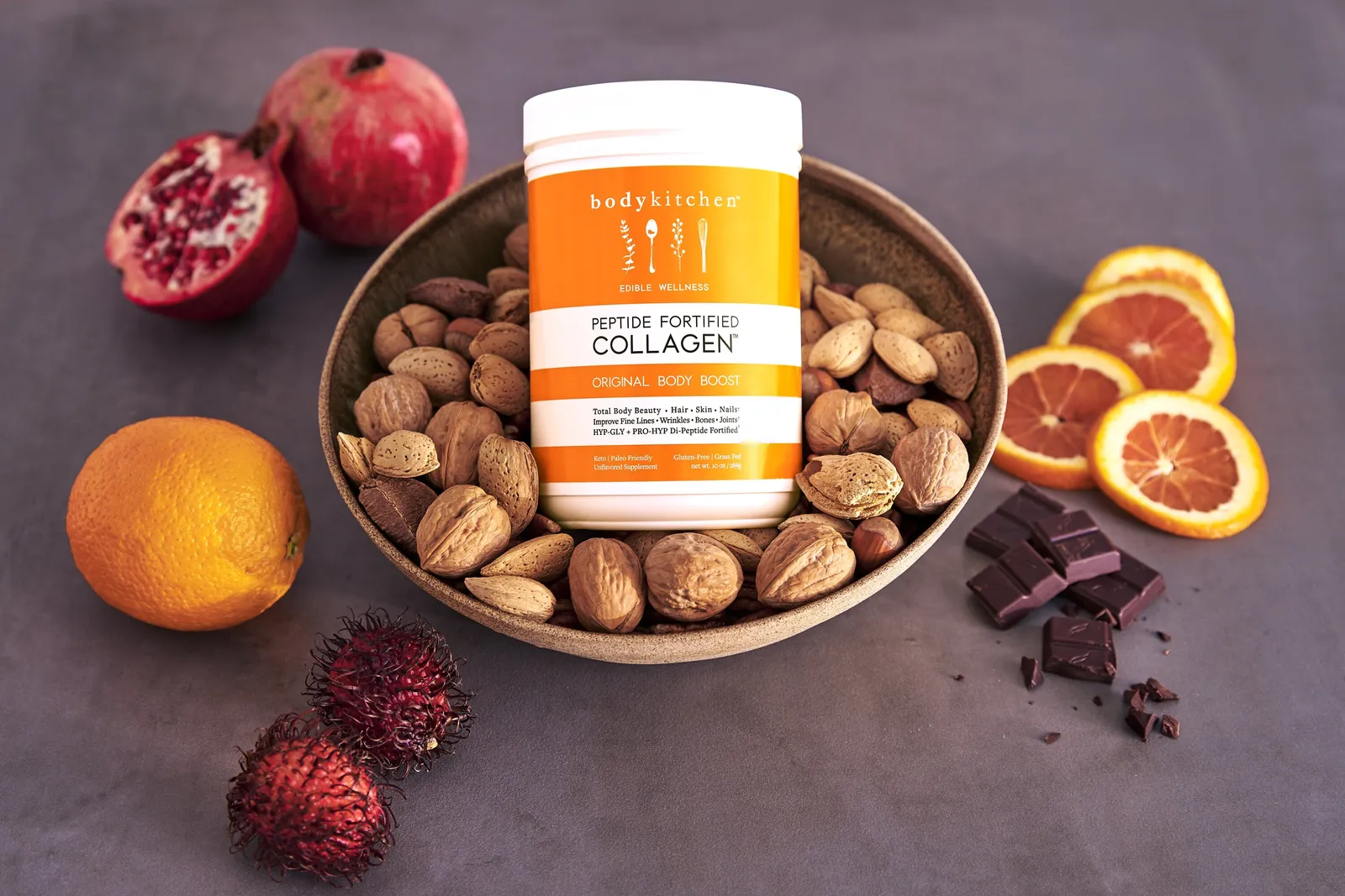 orange collagen jar nested in a bowl of almonds and walnuts, ringed by a cut pomegranate, blood oranges, chocolate pieces and rambutan on a gray surface