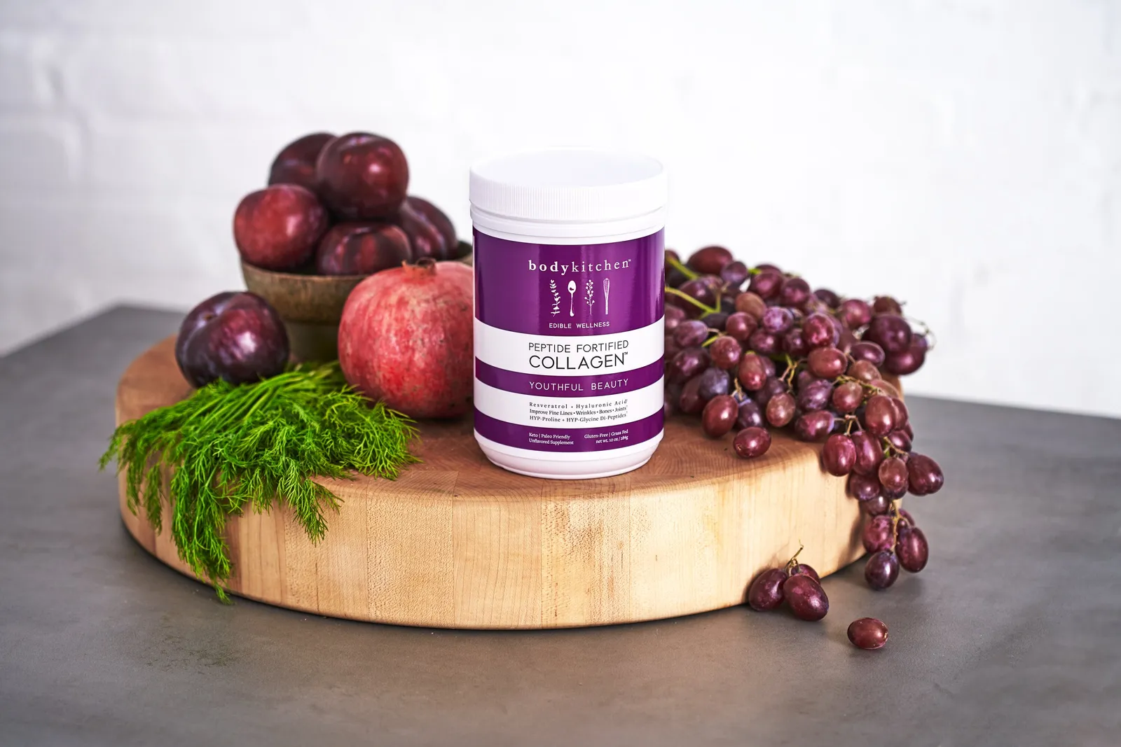 purple collagen jar on a round butcher block with plums, a pomegranate, dill and red grapes, on a gray counter before a white wall