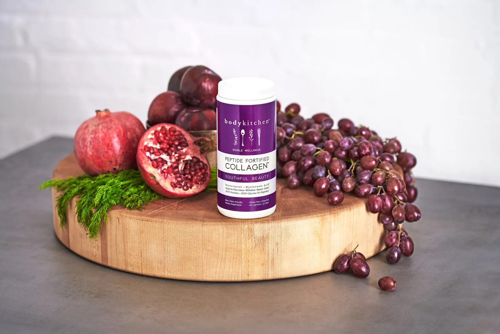 purple collagen jar on the same butcher block, now with a halved pomegranate showing its seeds beside dill, plums and red grapes