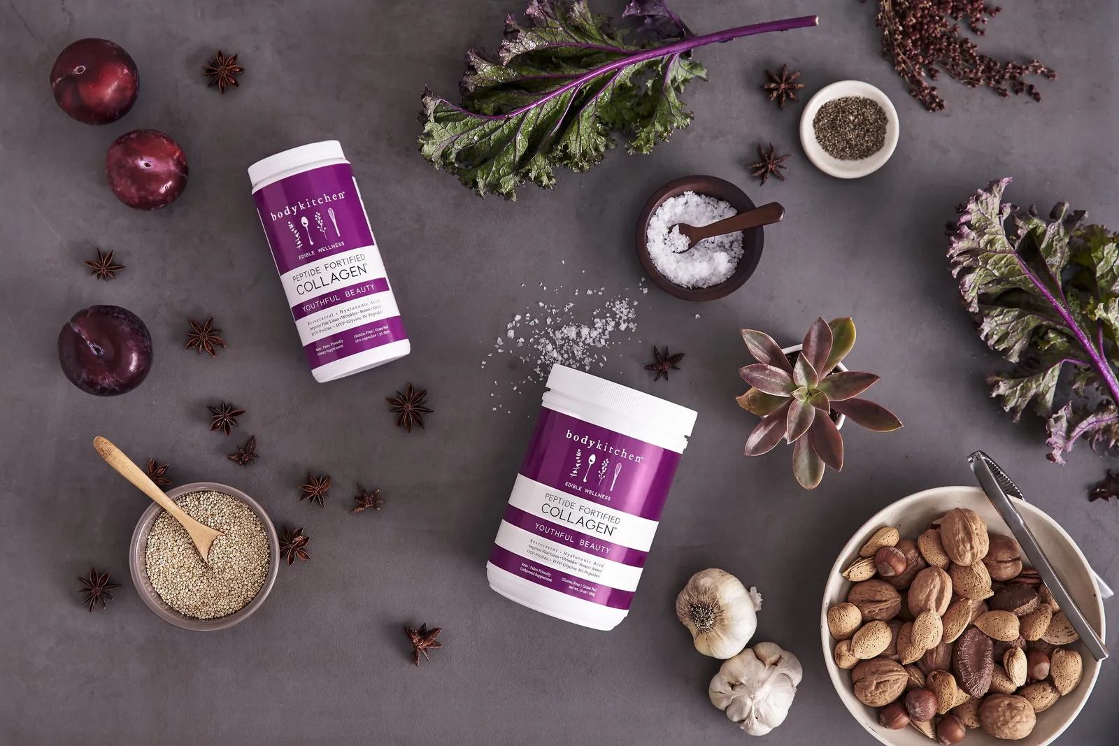 overhead view of two purple collagen jars on a gray surface among plums, kale, star anise, garlic, salt, grains and a bowl of nuts