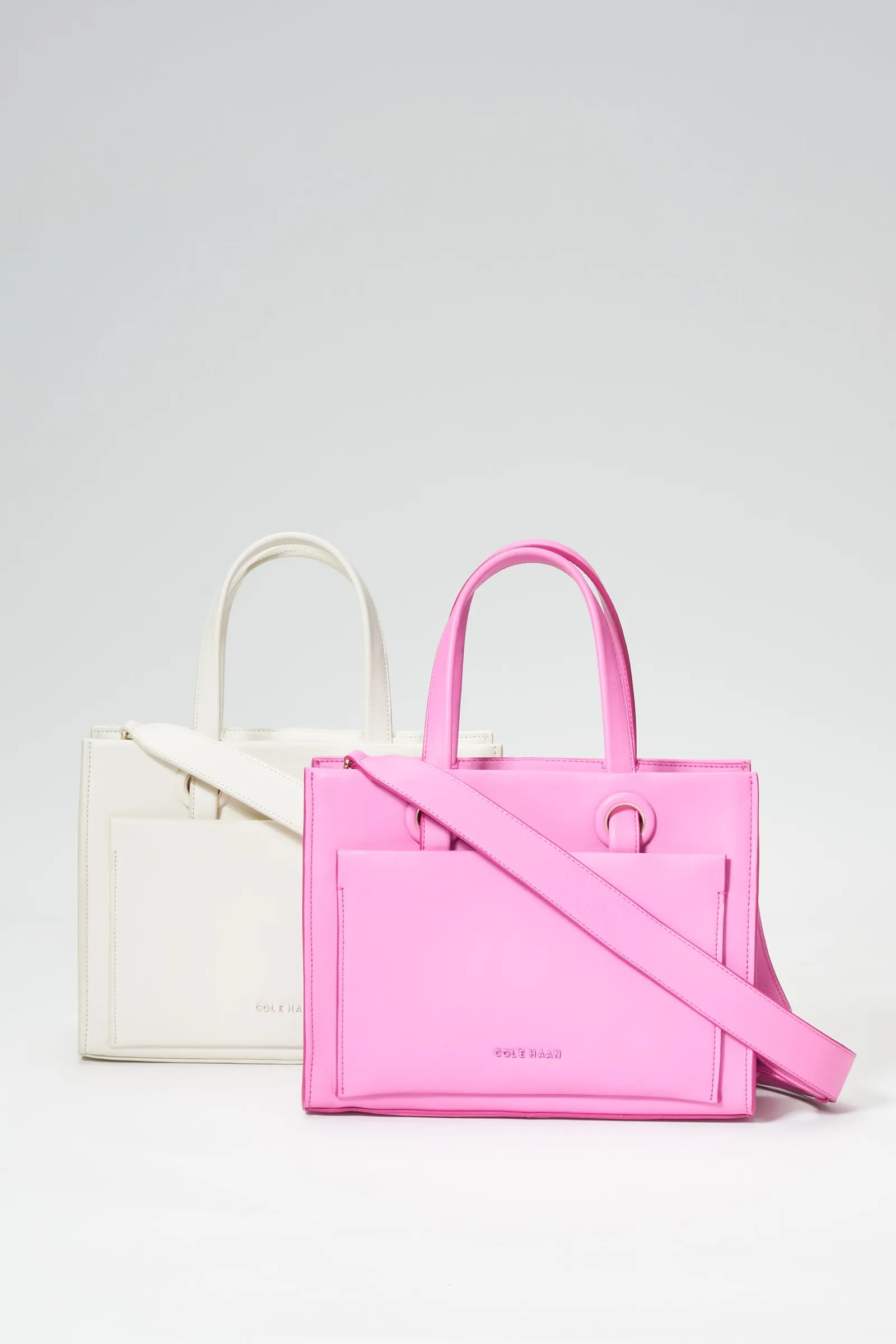 a pink structured tote with a long strap in front of a smaller cream tote, on a soft gray studio backdrop