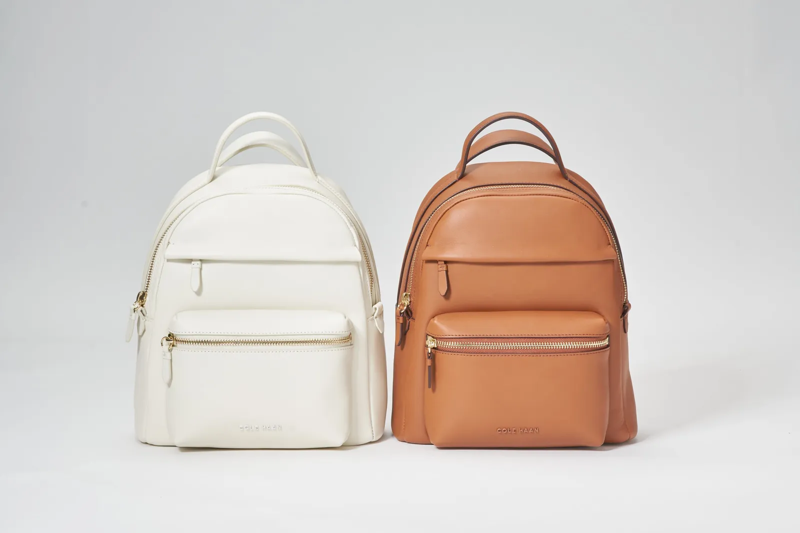 a cream leather backpack and a tan leather backpack standing side by side against a gray backdrop