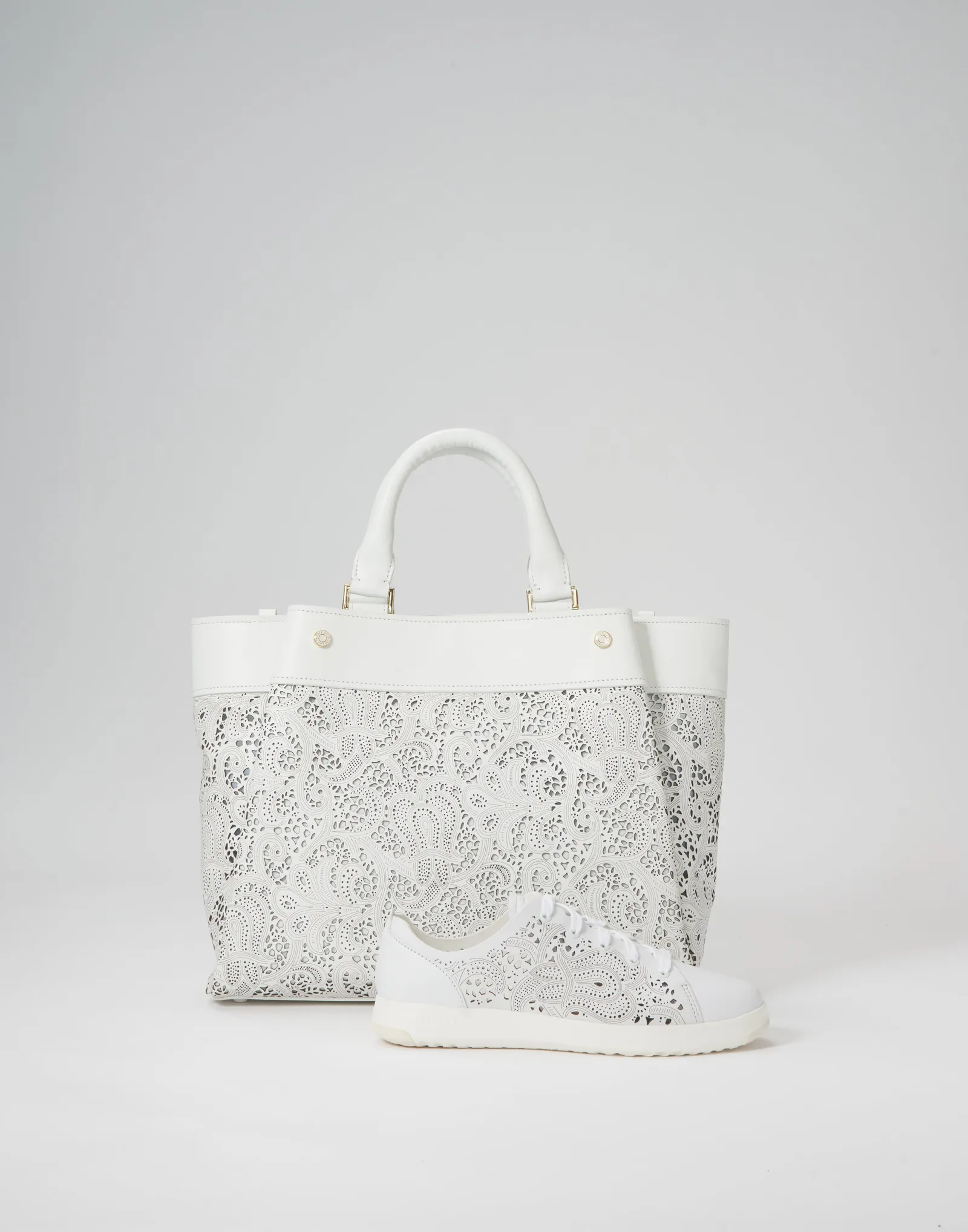 a white tote with laser-cut lace panels and a matching perforated white sneaker in front, on gray