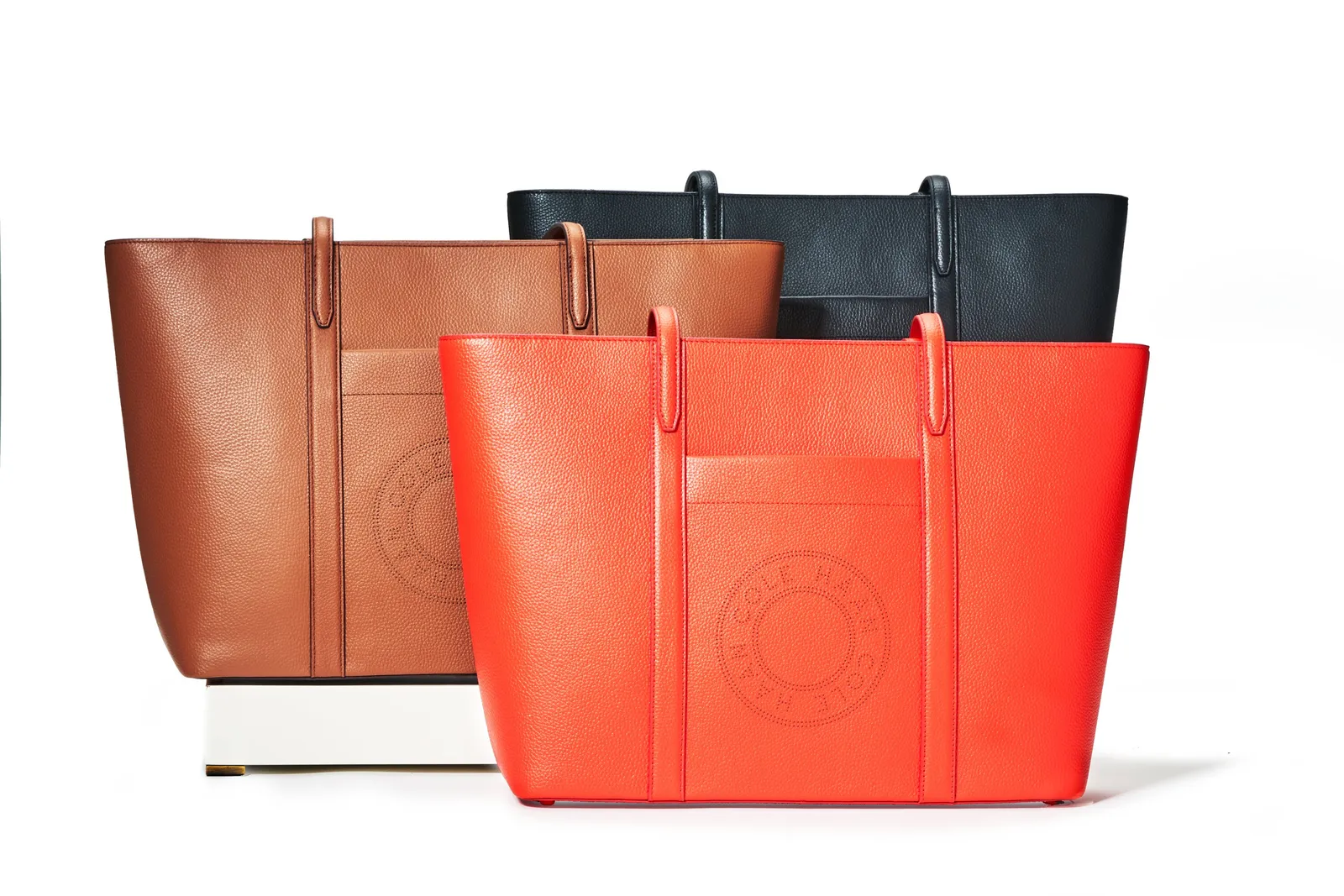 a tan tote, a black tote and a bright orange-red tote with an embossed circular logo, staggered on a white ledge