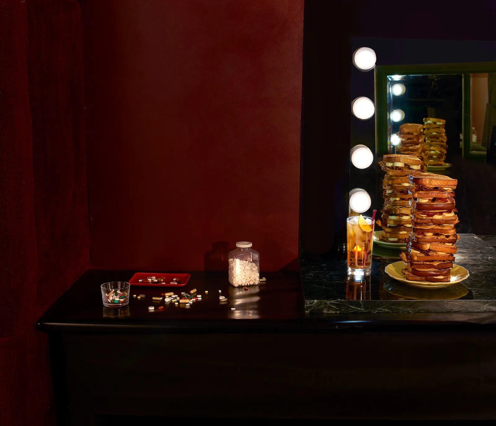 tall stacks of fried sandwiches on a mirrored dressing table with bulb lights, beside an iced tea, scattered pills and a pill bottle, against red curtains