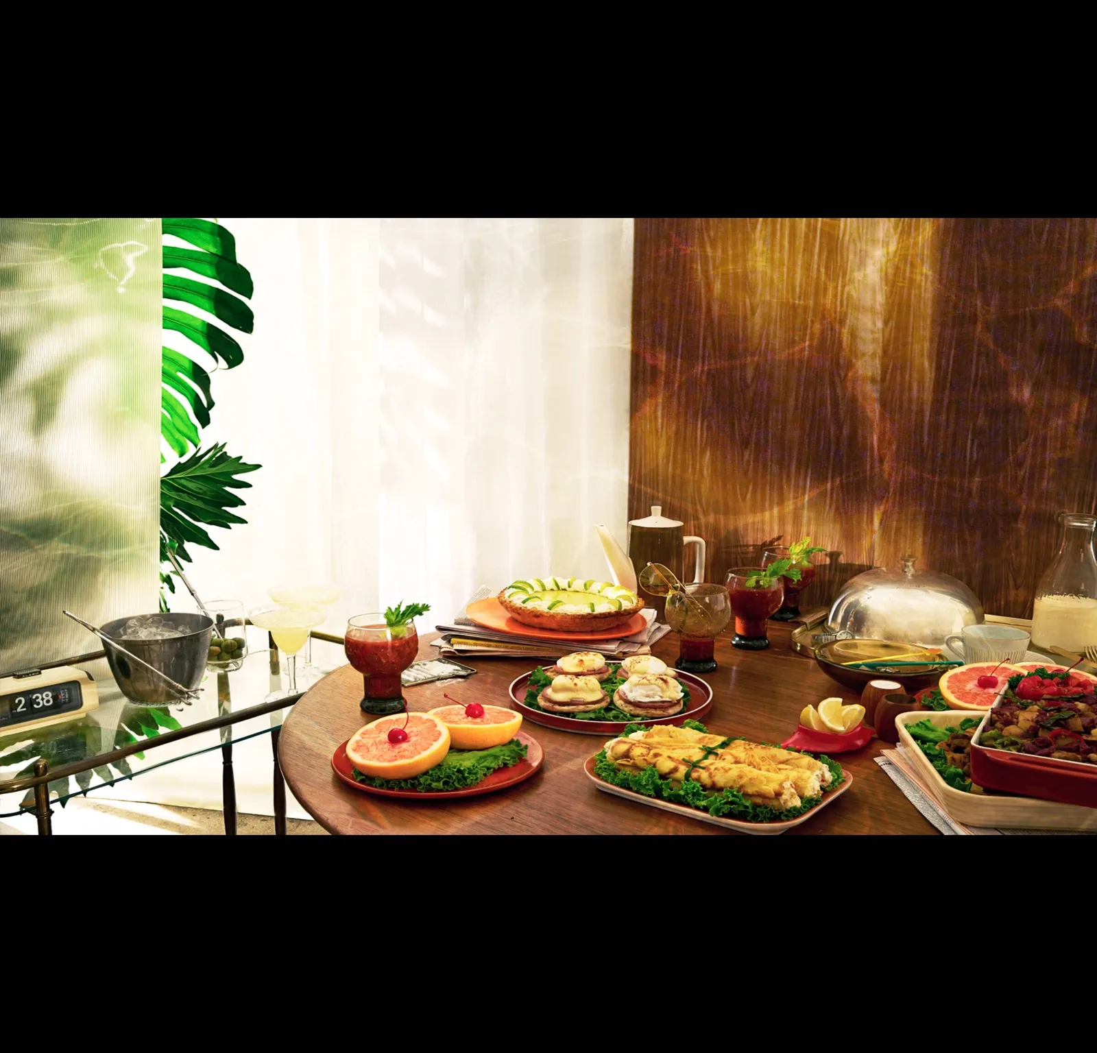 round wood table of retro brunch dishes, including grapefruit halves, eggs, crepes, cocktails and a pie, beside a glass cart and palm leaves in a sunlit room