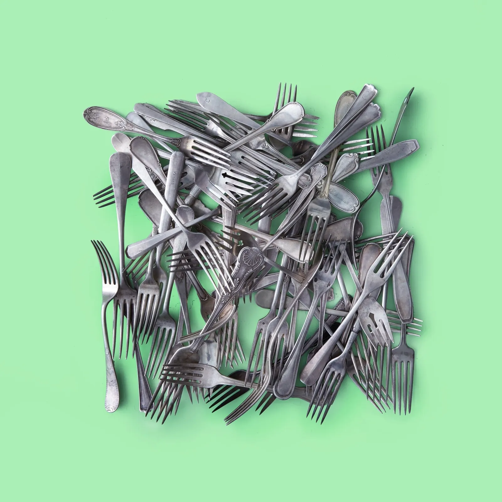 pile of tarnished silver forks in assorted patterns heaped into a square on a mint green background