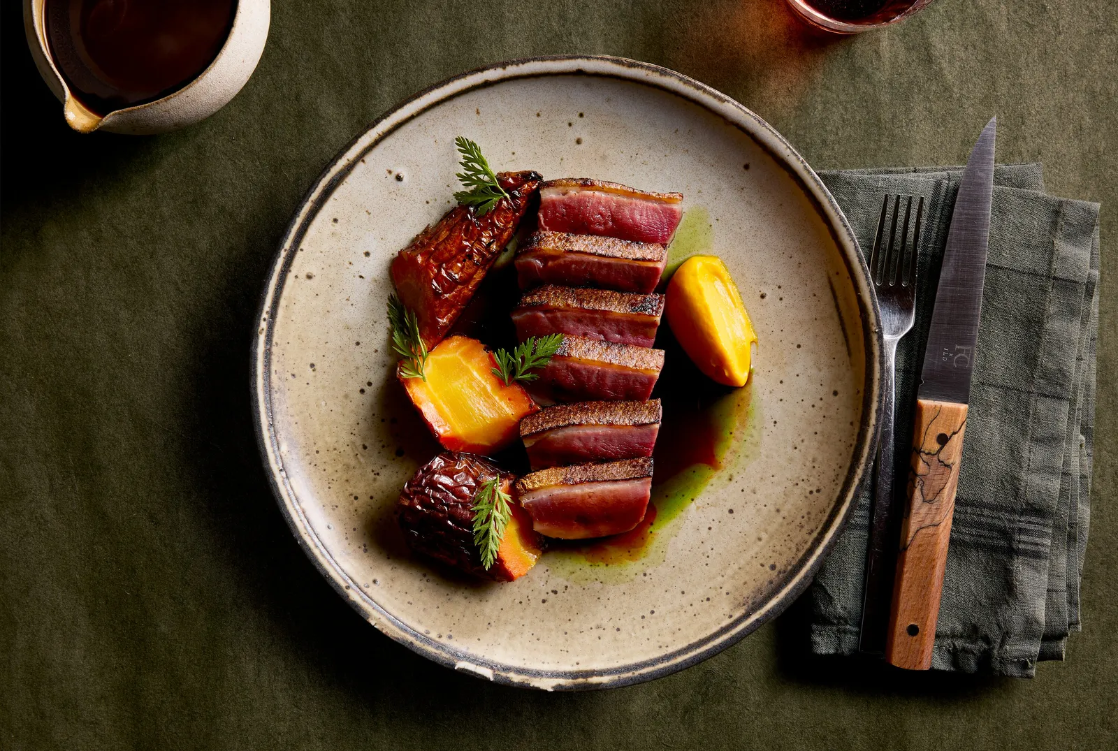 overhead view of sliced duck breast with stone fruit and herbs on a speckled plate, on olive green linen with a steak knife and sauce pitcher